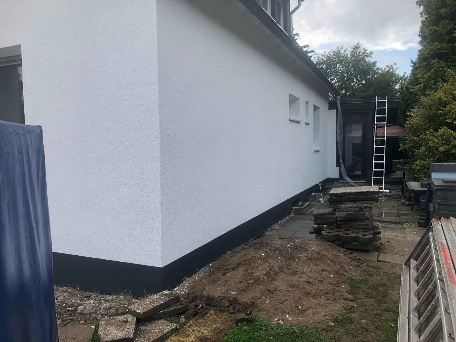 Świeżo pomalowana, biała elewacja domu w Przemyślu, z czarnym cokołem, widok z boku, z fragmentem trawnika i stertą płyt chodnikowych. W tle drabina oparta o budynek.