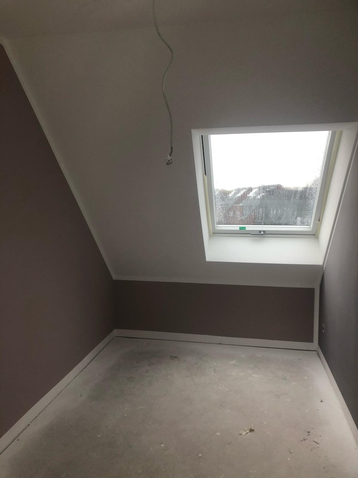 Pokój na poddaszu z oknem dachowym, ściany pomalowane w dwóch odcieniach brązu, surowa podłoga, widoczne kable zwisające z sufitu.