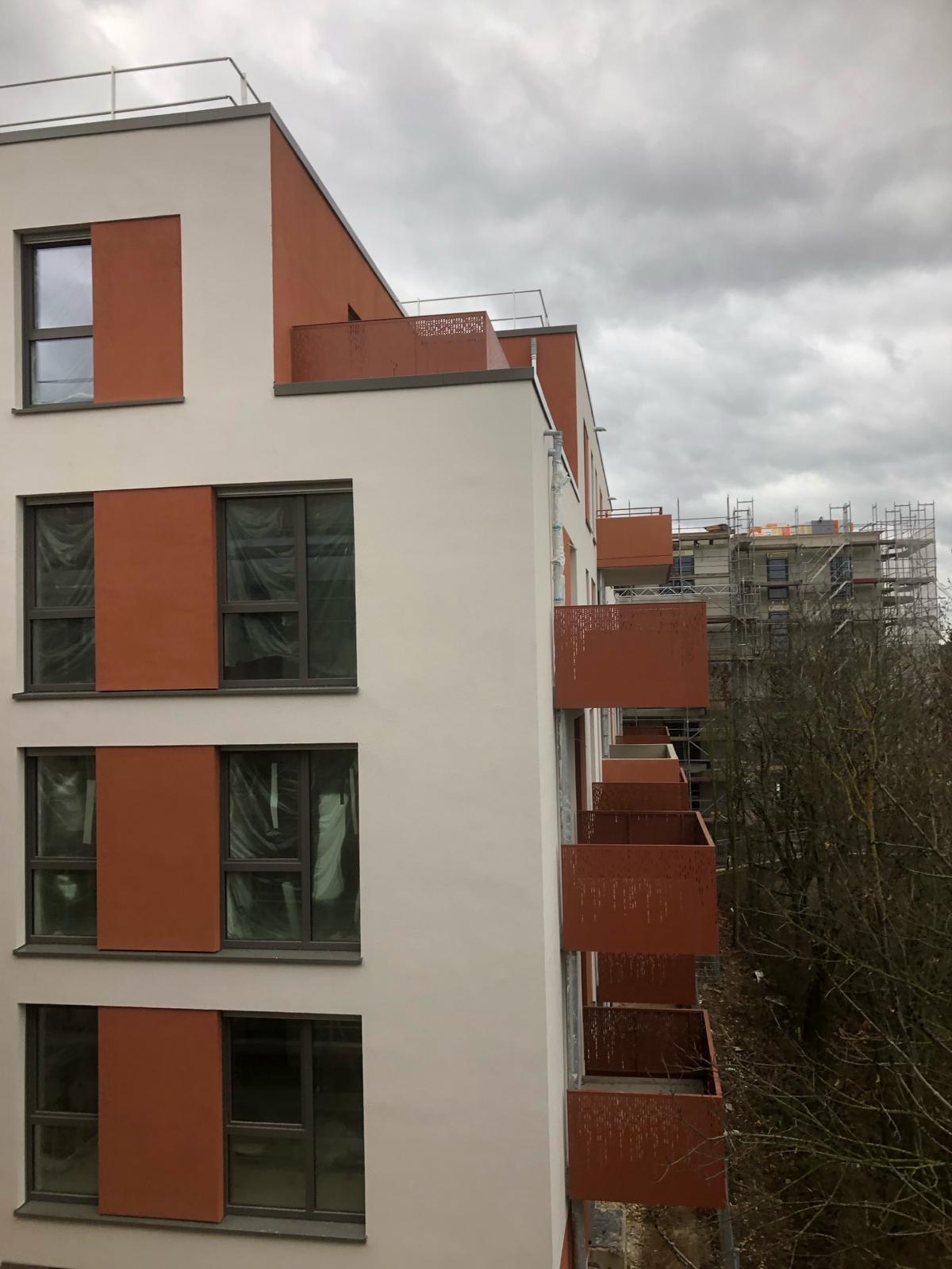 Nowoczesny budynek wielorodzinny z balkonami o rdzawym odcieniu, okna zabezpieczone folią, widoczne rusztowania na sąsiednim budynku w tle, pochmurne niebo.