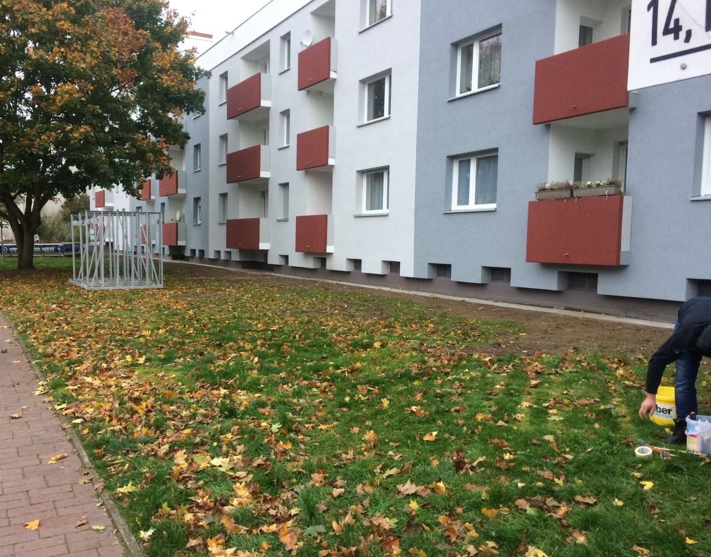 Fragment pomalowanego bloku mieszkalnego z czerwonymi balkonami, widoczna osoba przygotowująca farbę i narzędzia malarskie na trawniku pokrytym jesiennymi liśćmi.