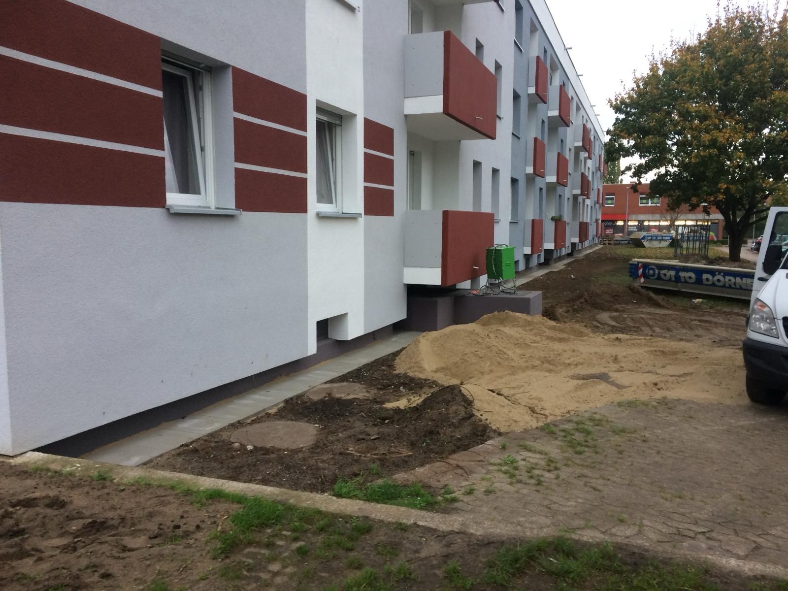 Budynek wielorodzinny z odnowioną elewacją w kolorach szarości, bieli i brązu, widoczne przygotowania terenu i sprzęt przed budynkiem.