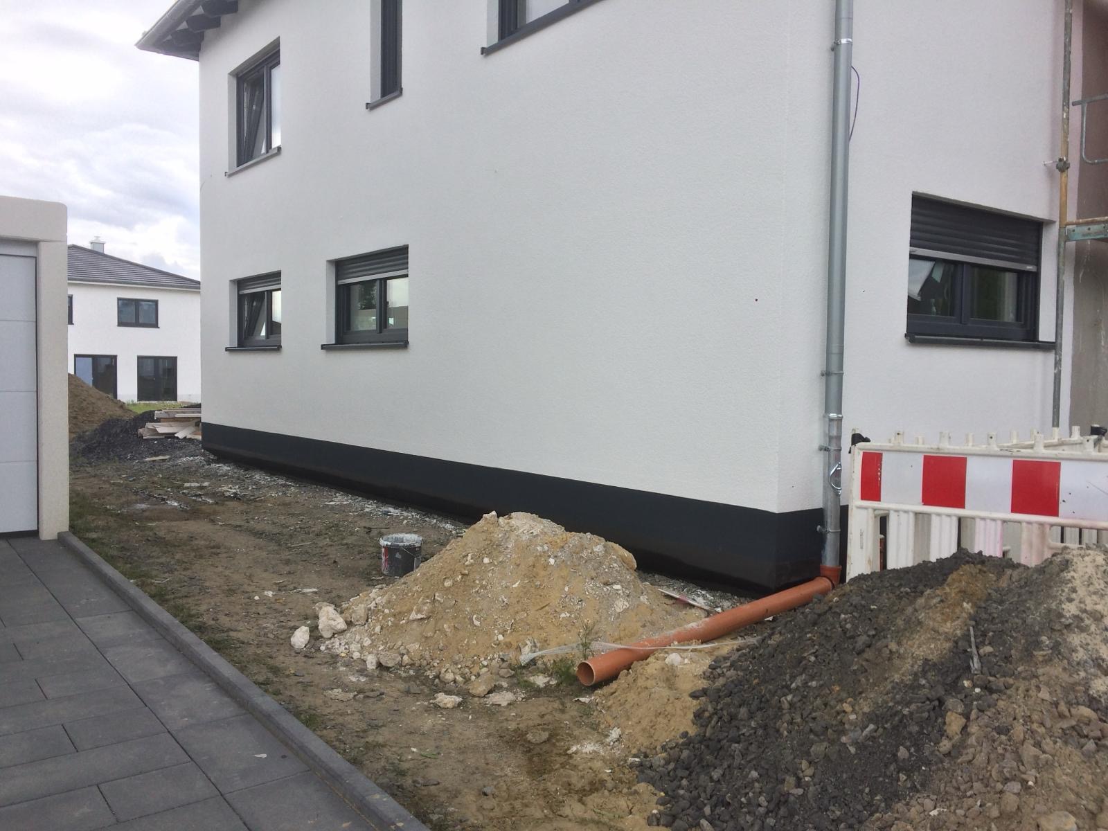 Świeżo otynkowany budynek jednorodzinny z szarą podmurówką i grafitowymi oknami, widoczny w trakcie prac ziemnych, z hałdami piasku i rurą kanalizacyjną przy elewacji.