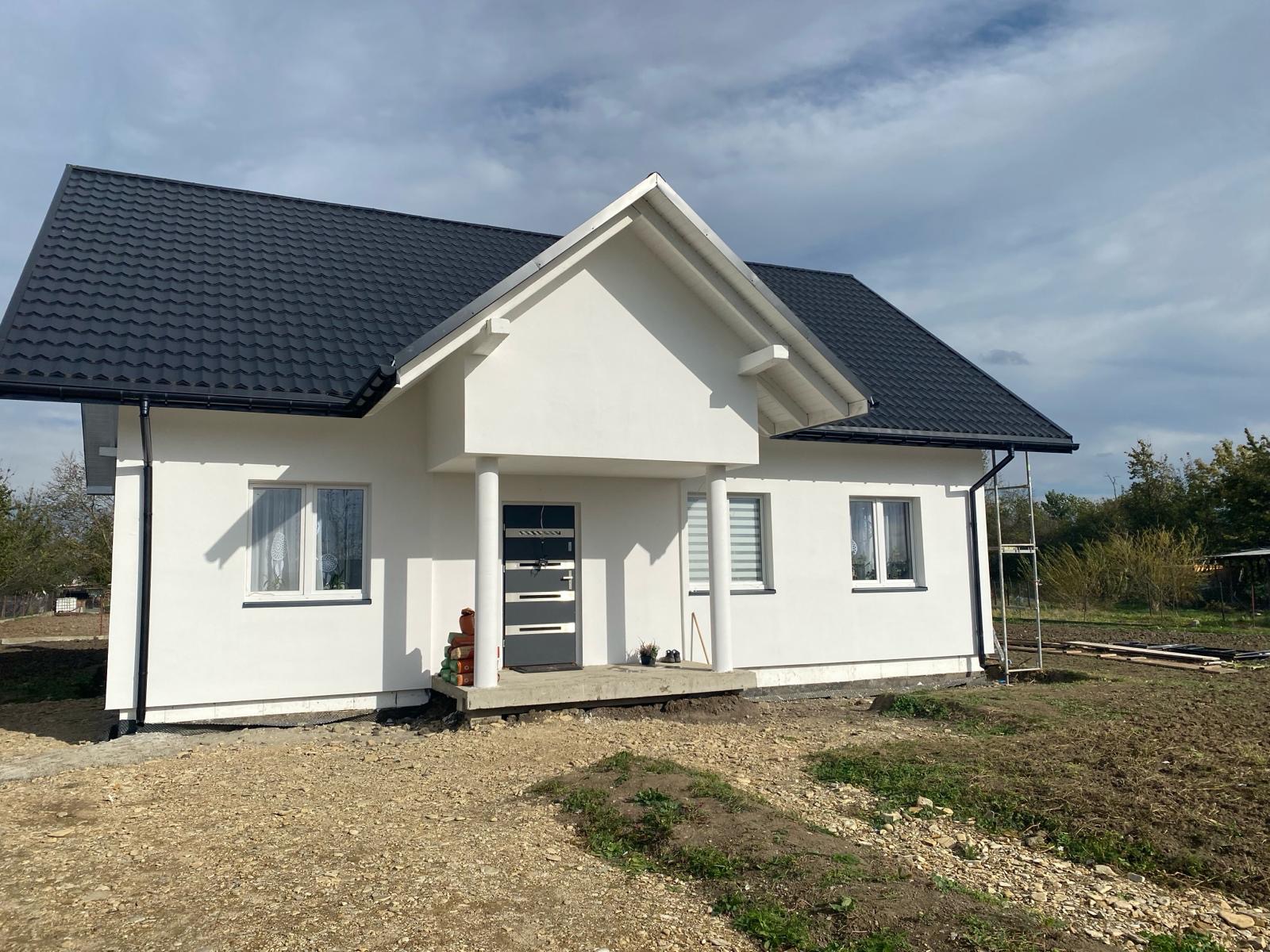 Nowoczesny dom jednorodzinny z białą elewacją, ciemnym dachem i grafitowymi elementami wykończeniowymi, widoczny w trakcie prac wykończeniowych wokół budynku.