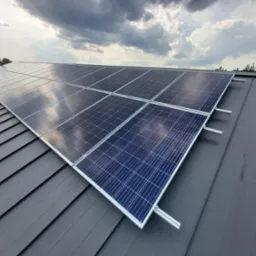 Panele fotowoltaiczne zamontowane na szarym, blaszany dachu, widok z góry na tle pochmurnego nieba.