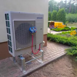 Klimatyzator Samsung na metalowym stelażu, podłączony do manometru serwisowego z czerwonymi przewodami, na tle brukowanego chodnika i zadbanego ogrodu z ż&oacute;łtym samochodem serwisowym w oddali.