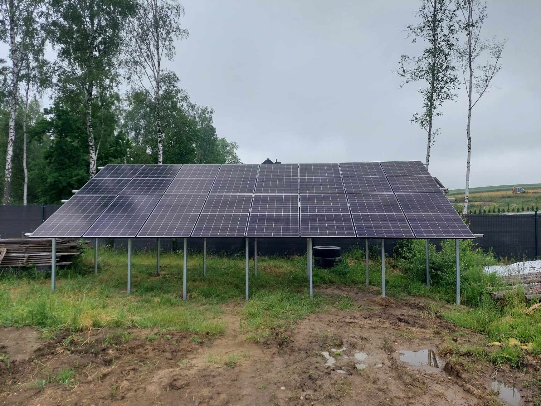 Instalacja paneli słonecznych na metalowych podporach, widok na polne tło i pochmurne niebo.