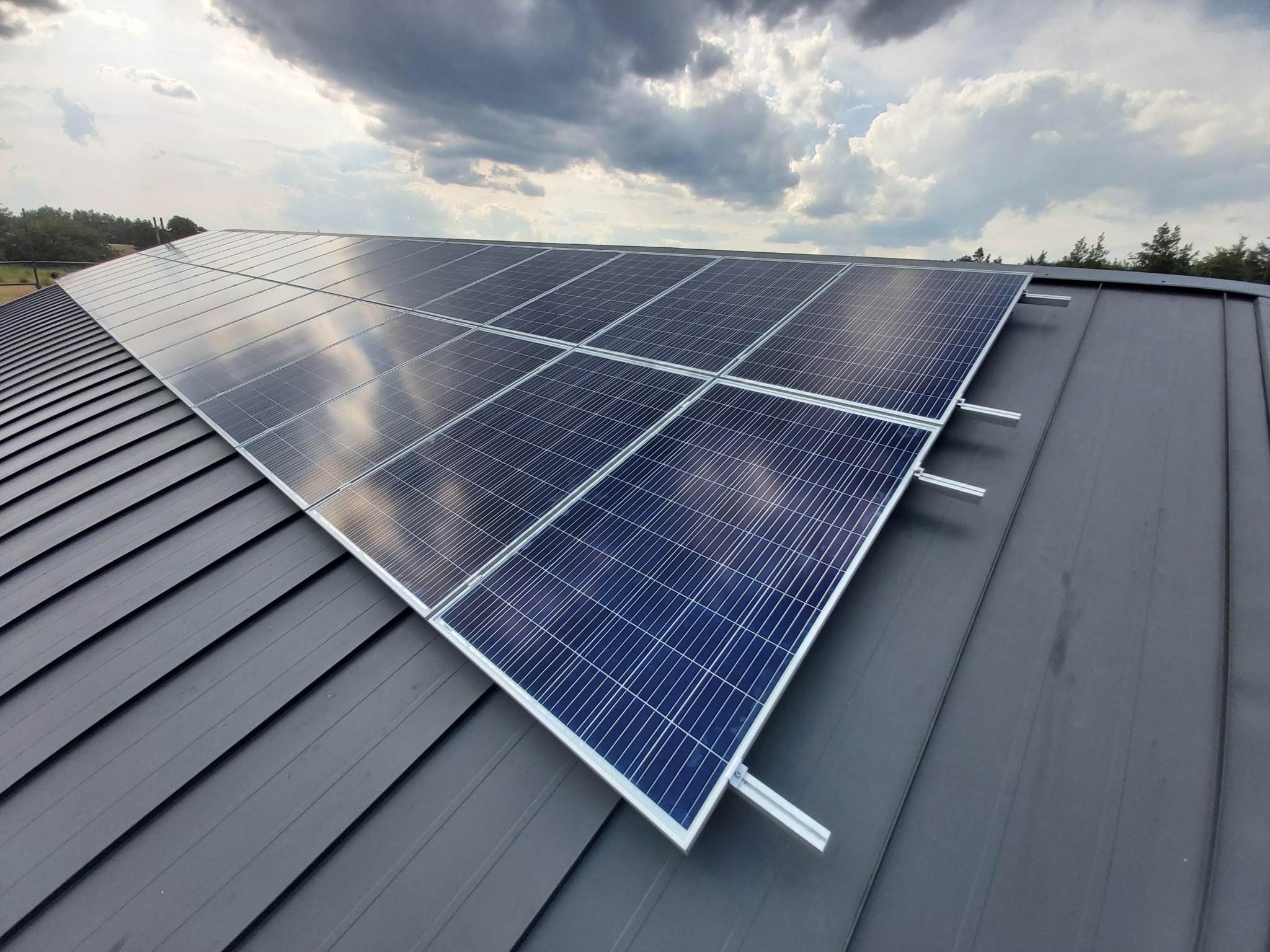 Panele fotowoltaiczne zamontowane na szarym, blaszany dachu, widok z góry na tle pochmurnego nieba.