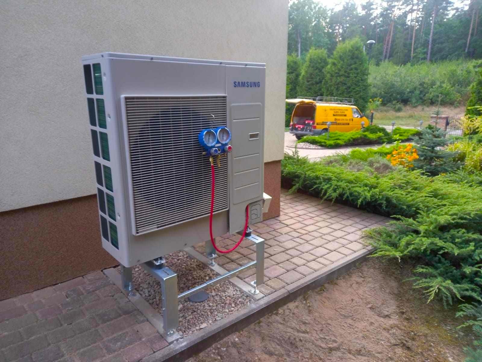 Klimatyzator Samsung na metalowym stelażu, podłączony do manometru serwisowego z czerwonymi przewodami, na tle brukowanego chodnika i zadbanego ogrodu z żółtym samochodem serwisowym w oddali.