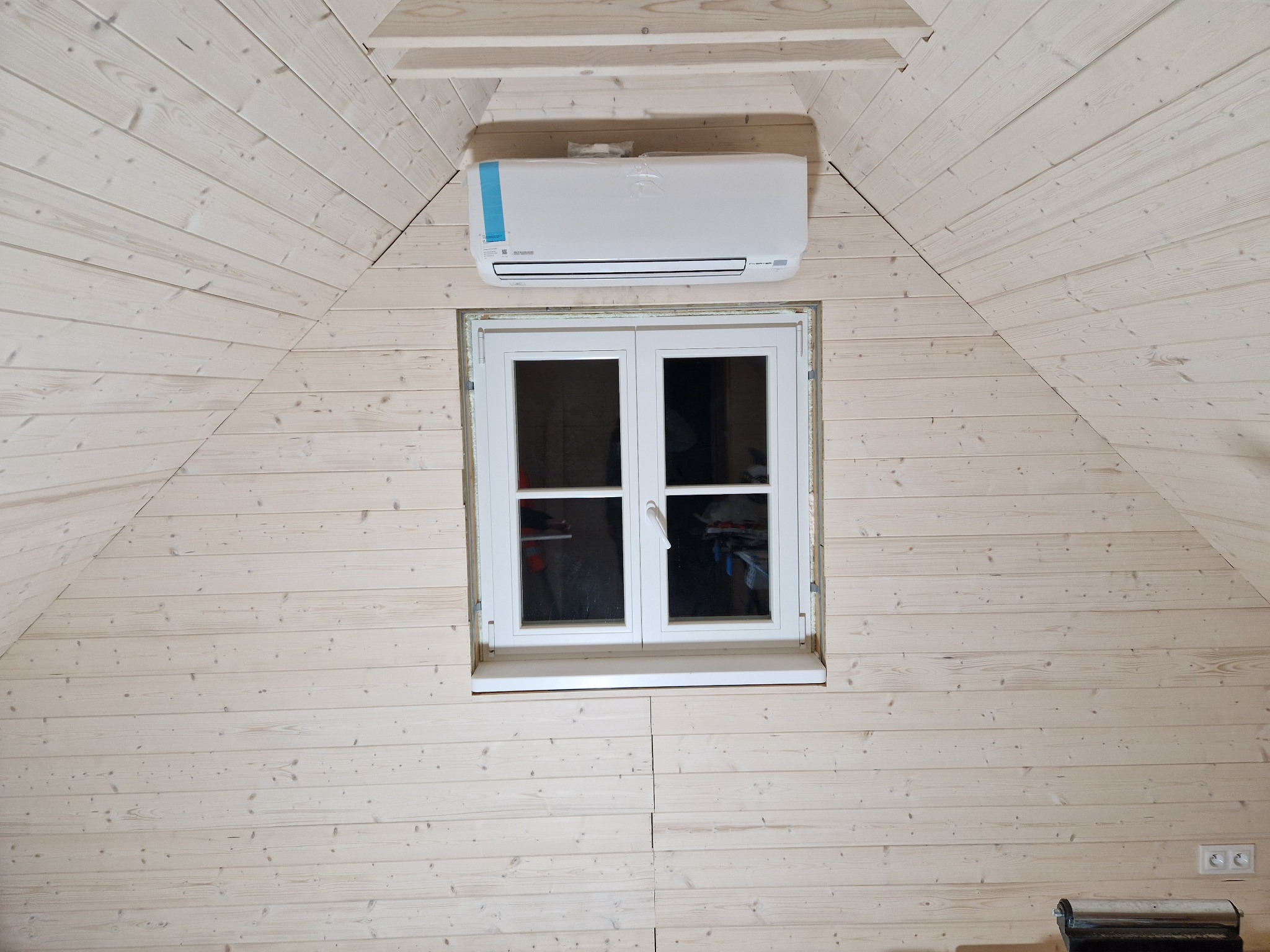 Klimatyzator ścienny zamontowany nad oknem w pomieszczeniu wykończonym jasną boazerią drewnianą, widoczne gniazdka elektryczne na ścianie.