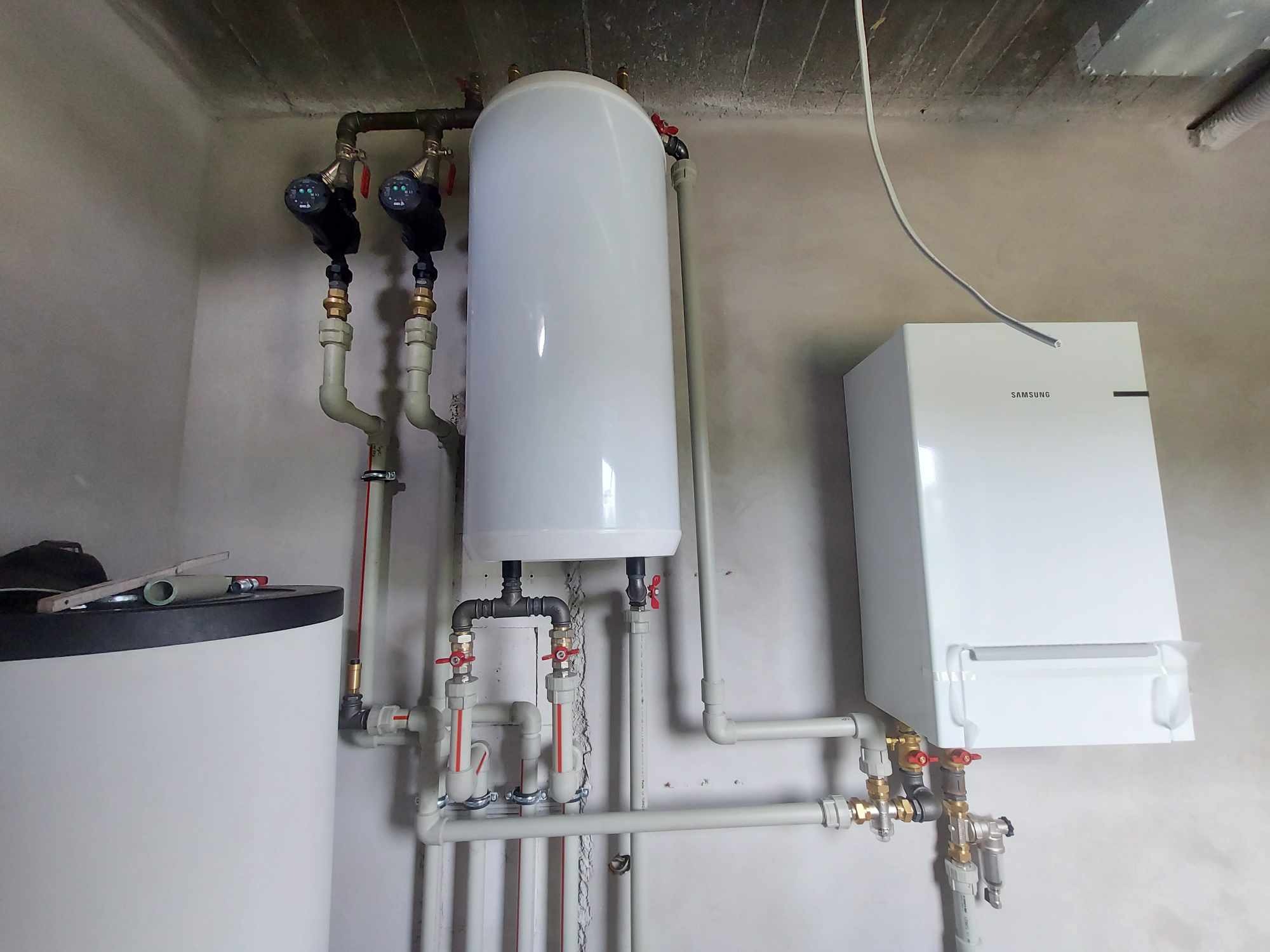 Biały, wiszący piec gazowy Samsung z podłączonymi rurami, obok pionowy zbiornik na wodę i dodatkowy, mniejszy zbiornik z widocznymi narzędziami na górze, całość instalacji w surowym pomieszczeniu.