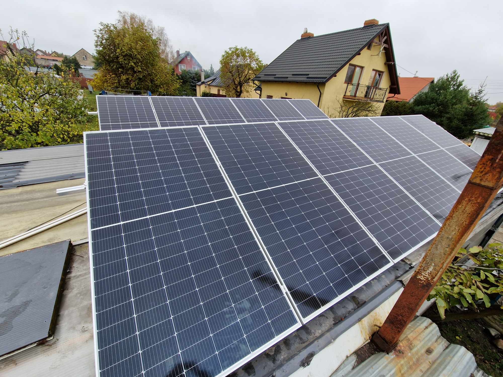 Zestaw paneli fotowoltaicznych na dachu budynku mieszkalnego, widoczne zardzewiałe elementy konstrukcyjne, pochmurne niebo w tle.