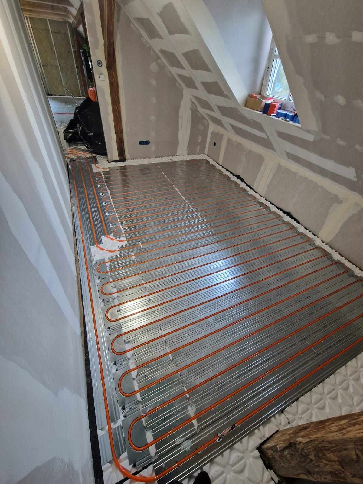 Podłogówka w trakcie montażu w pomieszczeniu na poddaszu, rury grzewcze ułożone na specjalnych płytach izolacyjnych, ściany w stanie surowym.
