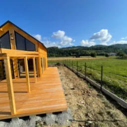 Drewniany dom z tarasem w budowie, widok na zielone wzgórza i niebo. W tle częściowo ukończone ogrodzenie panelowe. Całość w słoneczny dzień.