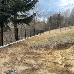 Łukasz Szlembarski - Nowo postawione, czarne ogrodzenie panelowe z betonową podmurówką na nierównym terenie. Ślady opon na ziemi. W tle drzewa i pochmurne niebo.