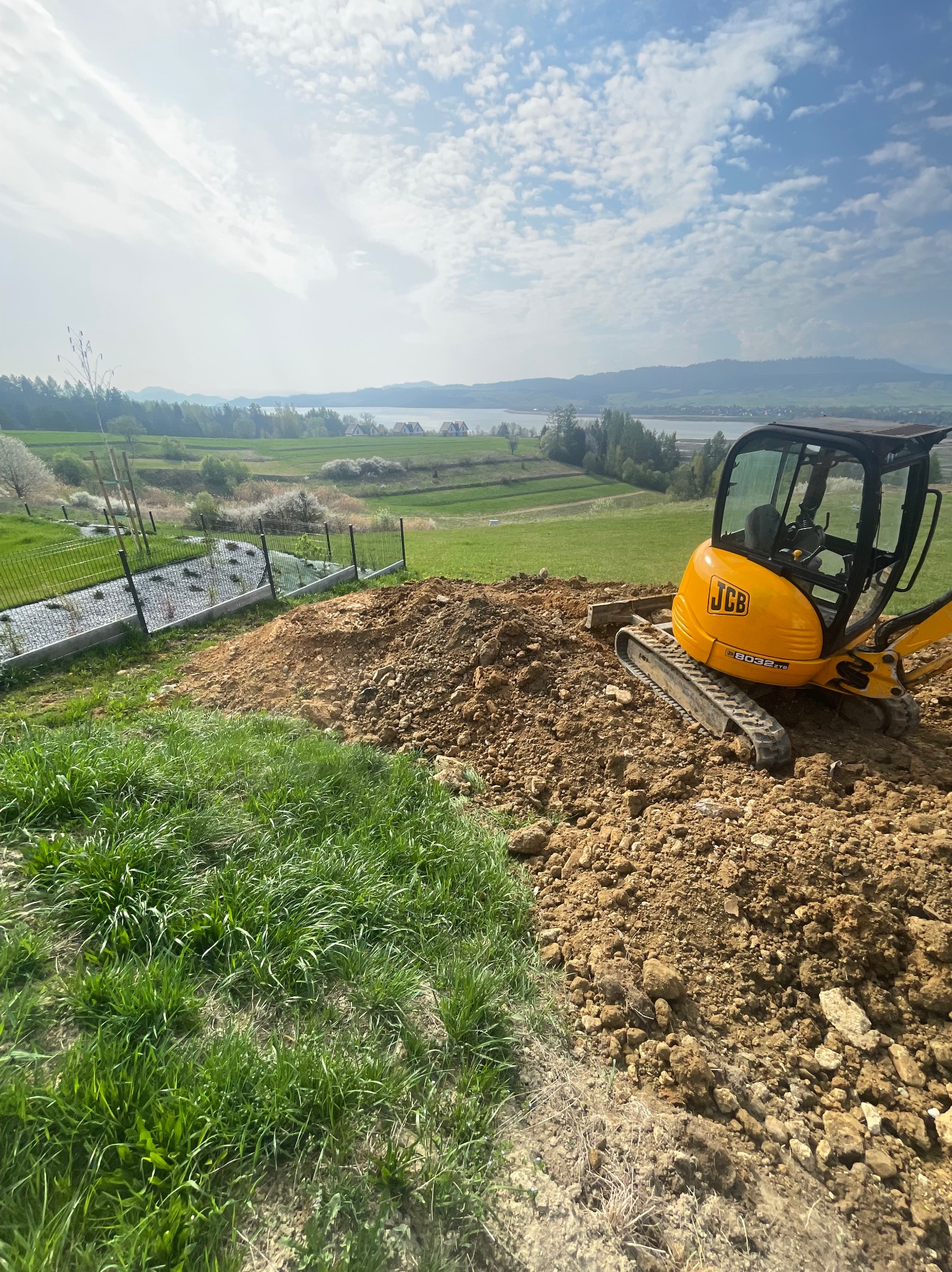 Żółta mini koparka JCB na wzgórzu z ziemią, w tle zielony krajobraz z jeziorem i ogrodzeniem panelowym. Niwelacja terenu w toku.