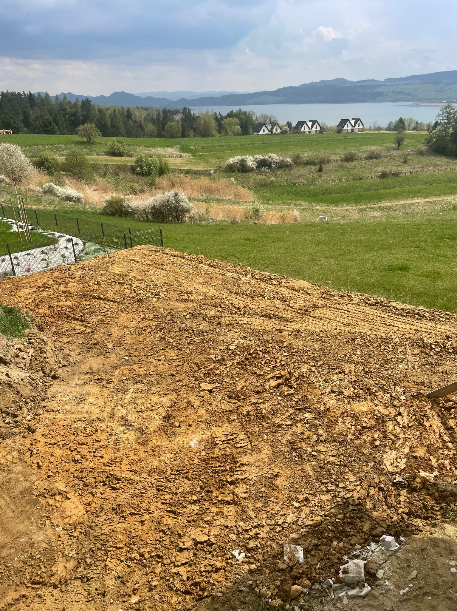 Wyrównany teren pod budowę z widokiem na krajobraz z jeziorem i domami w tle. Częściowo widoczne ogrodzenie panelowe.