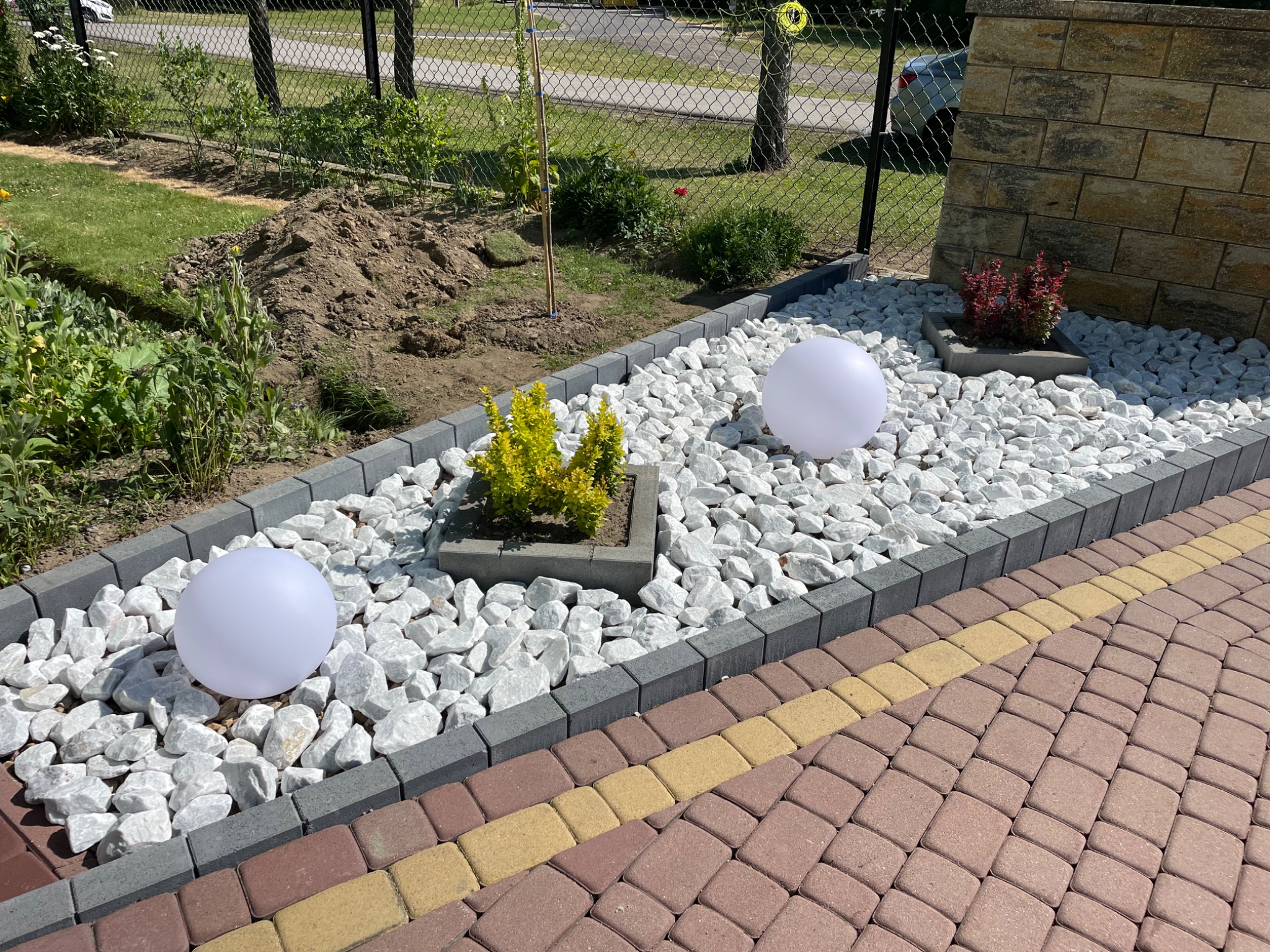 Ogród z białymi kamieniami, obrzeżami z kostki, roślinami w donicach i kulami świetlnymi, widok z góry na ścieżkę z kostki brukowej w dwóch kolorach.