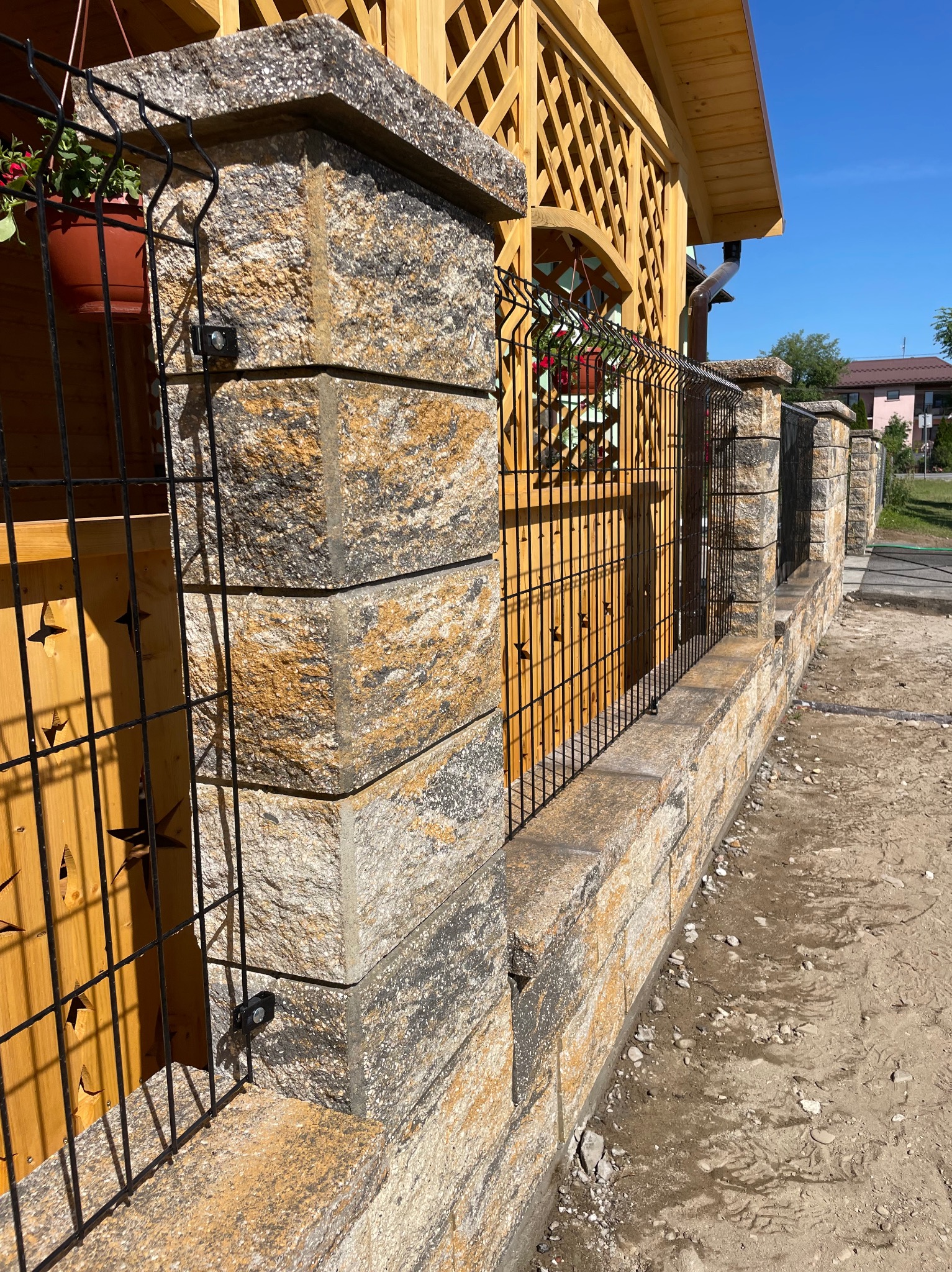 Nowoczesne ogrodzenie panelowe z kamiennymi słupkami i drewnianymi elementami dekoracyjnymi, częściowo zamontowane, w słoneczny dzień.