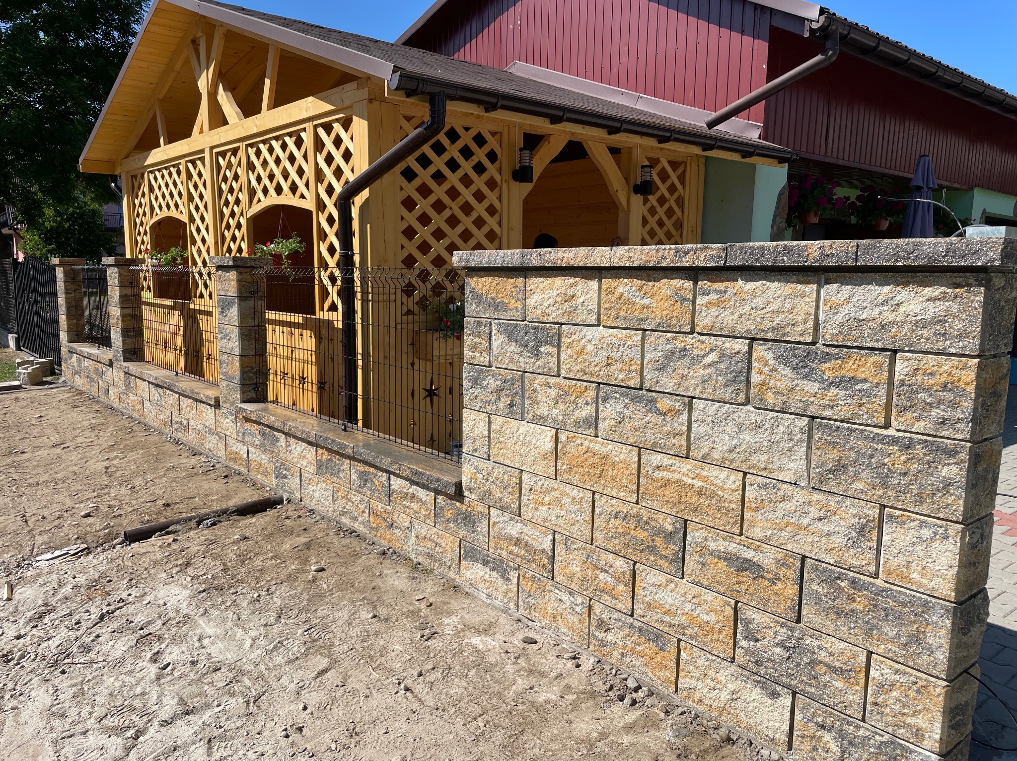 Kamienny mur z elementami ogrodzenia panelowego i drewnianą altaną w tle, w słoneczny dzień. Widoczne niedokończone prace ziemne.