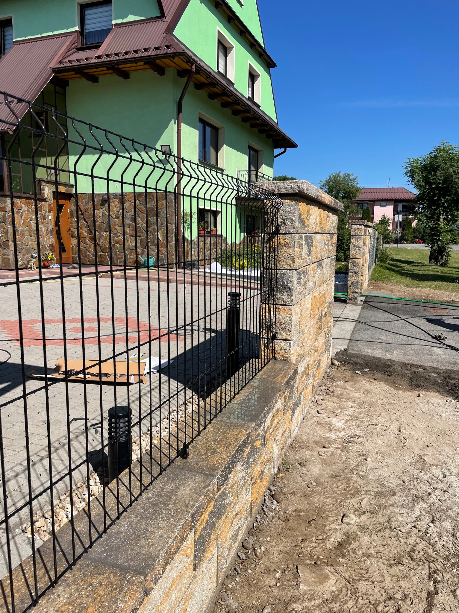 Montaż czarnego ogrodzenia panelowego na murku z kamienia piaskowca. Widoczne elementy montażowe i narzędzia. W tle zielony dom jednorodzinny i błękitne niebo.