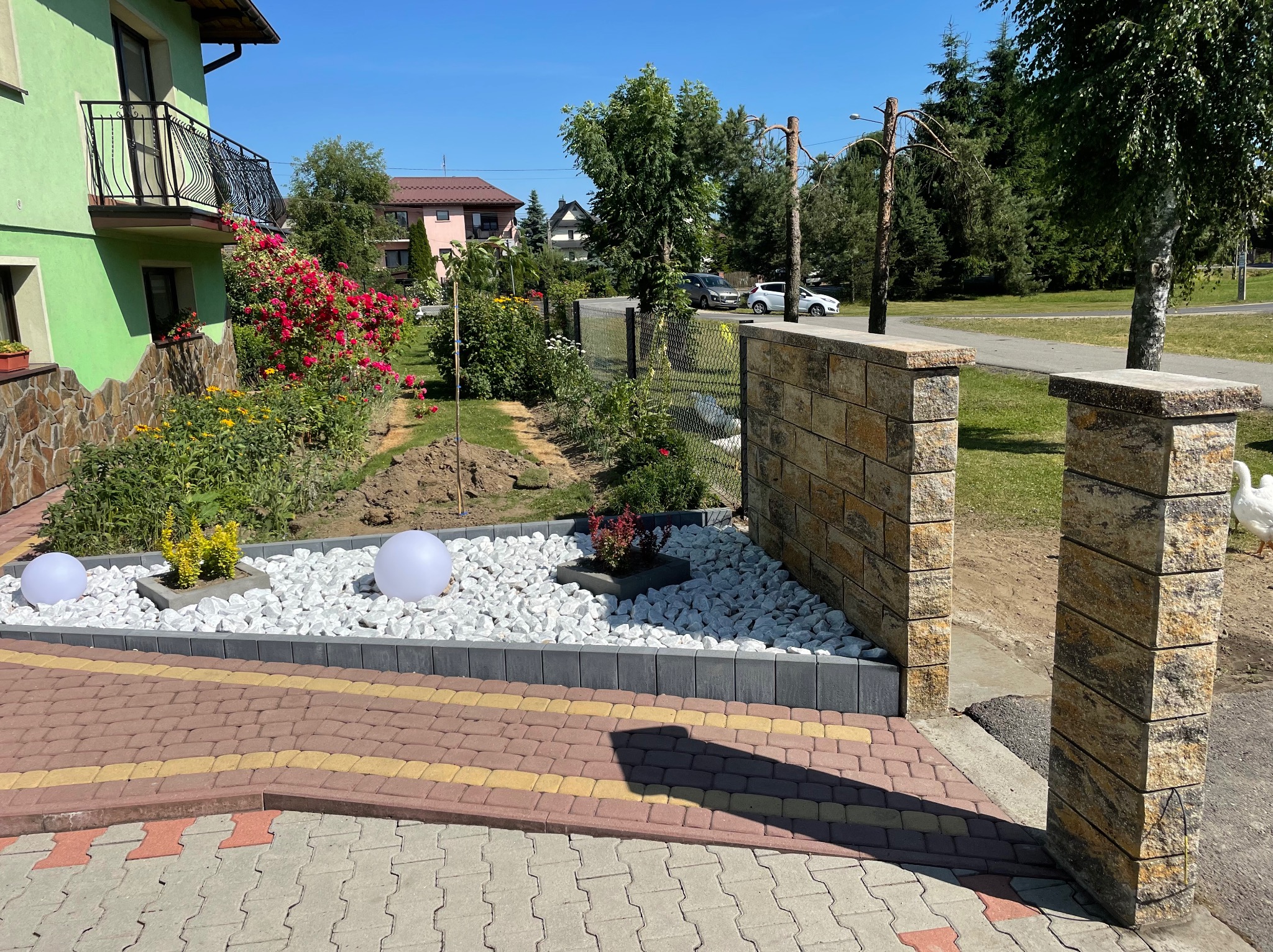 Ogród z dekoracyjnym murem z kamienia, białymi kamieniami i kulistymi lampami. Widoczny fragment domu z balkonem i kolorowa kostka brukowa na podjeździe.