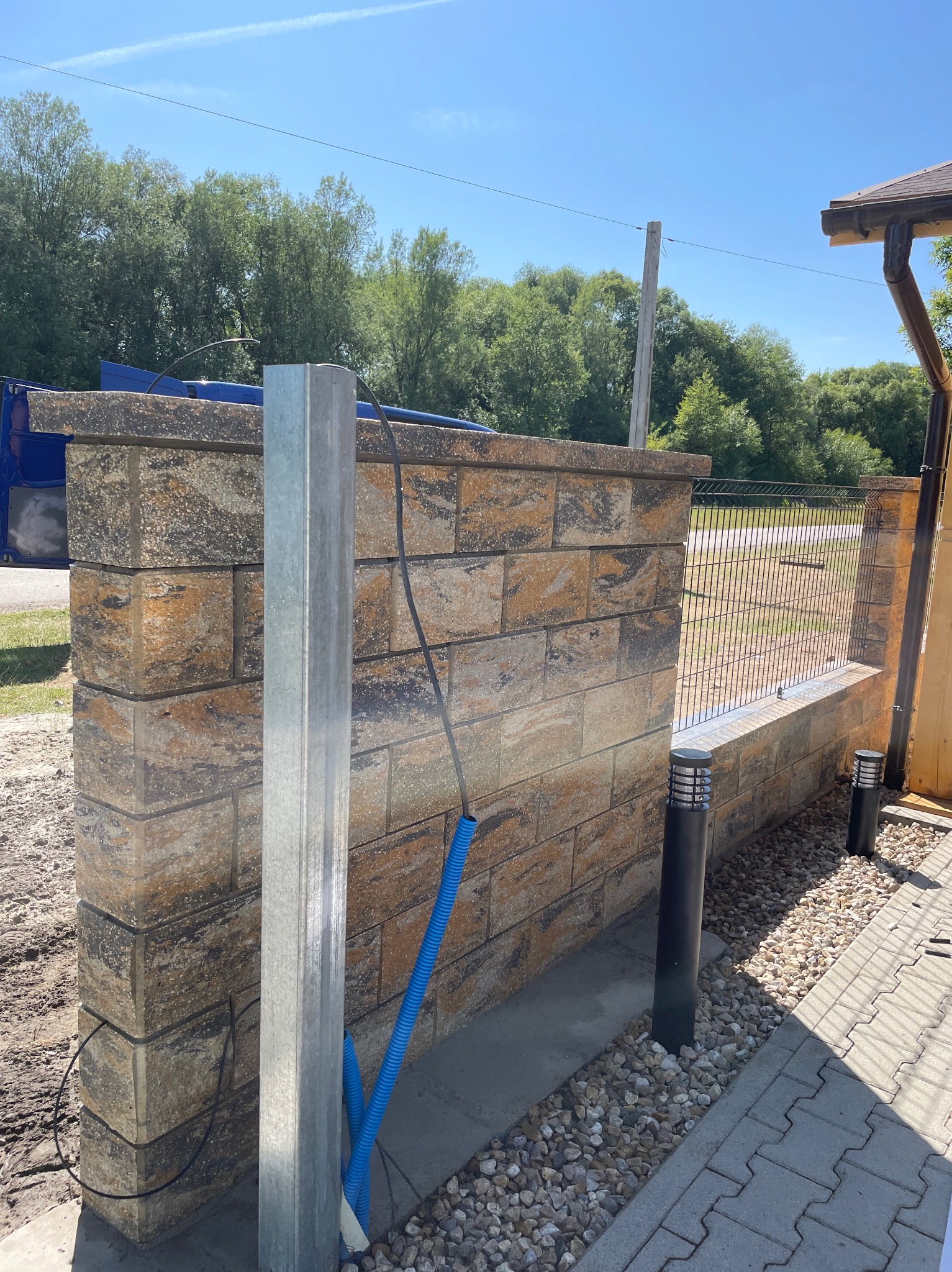 Ogrodzenie z paneli na murku z kamienia łupanego, słupki oświetleniowe i podsypka żwirowa. Widok na posesję przez ogrodzenie.