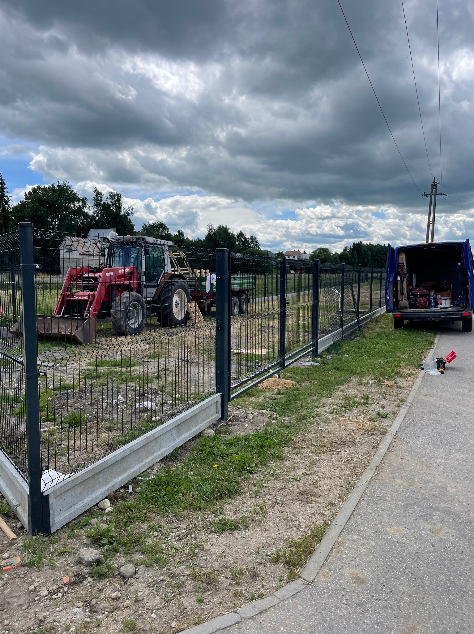 Montaż ogrodzenia panelowego z podmurówką betonową. W tle traktor i samochód dostawczy. Pochmurne niebo.