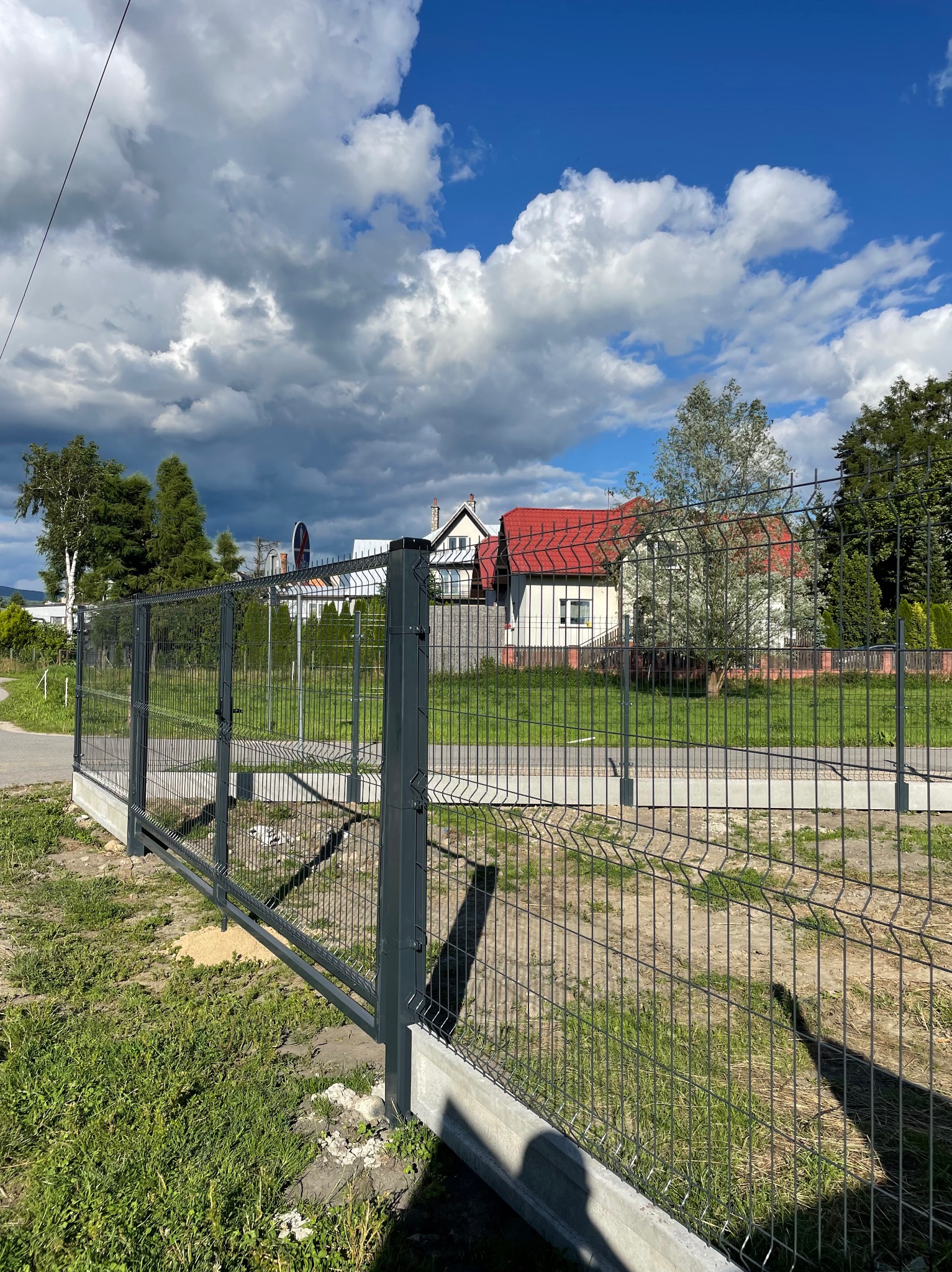 Nowoczesne, szare ogrodzenie panelowe na betonowej podmurówce, widok z zewnątrz posesji na tle domu z czerwonym dachem i błękitnego nieba z chmurami.