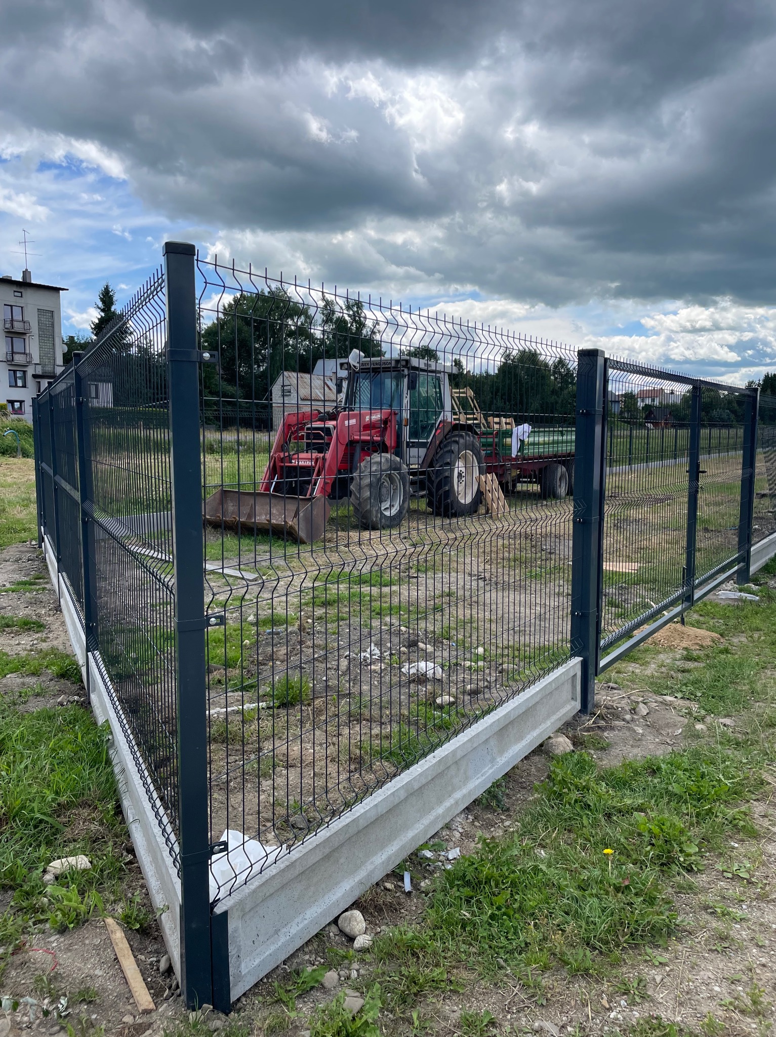 Nowoczesne ogrodzenie panelowe z podmurówką betonową, zza którego wyłania się czerwony ciągnik rolniczy z przyczepą na tle pochmurnego nieba.