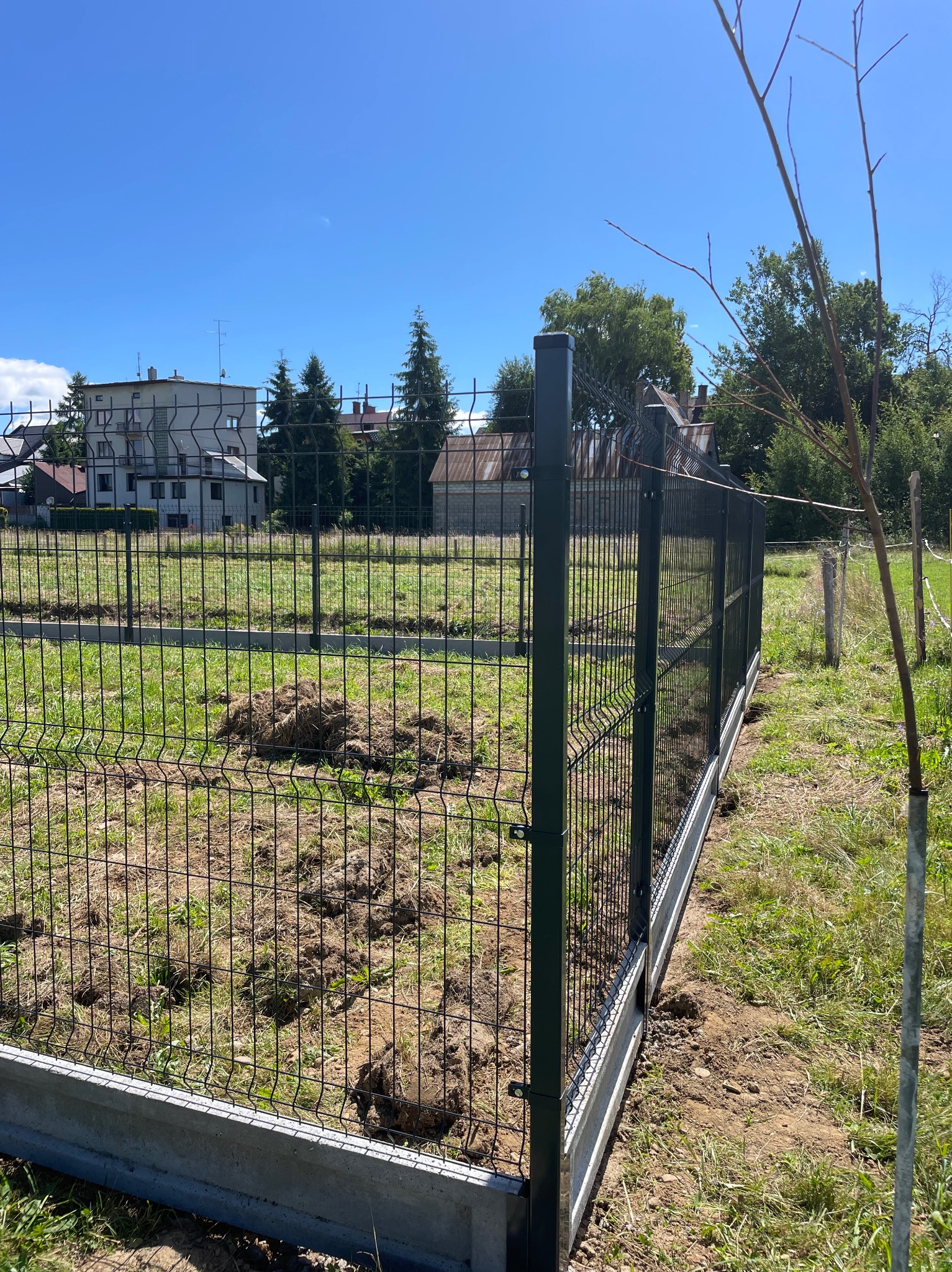 Nowoczesne ogrodzenie panelowe z podmurówką betonową na trawiastym terenie. W tle budynki mieszkalne i drzewa, błękitne niebo. Dbałość o detale wykończenia.
