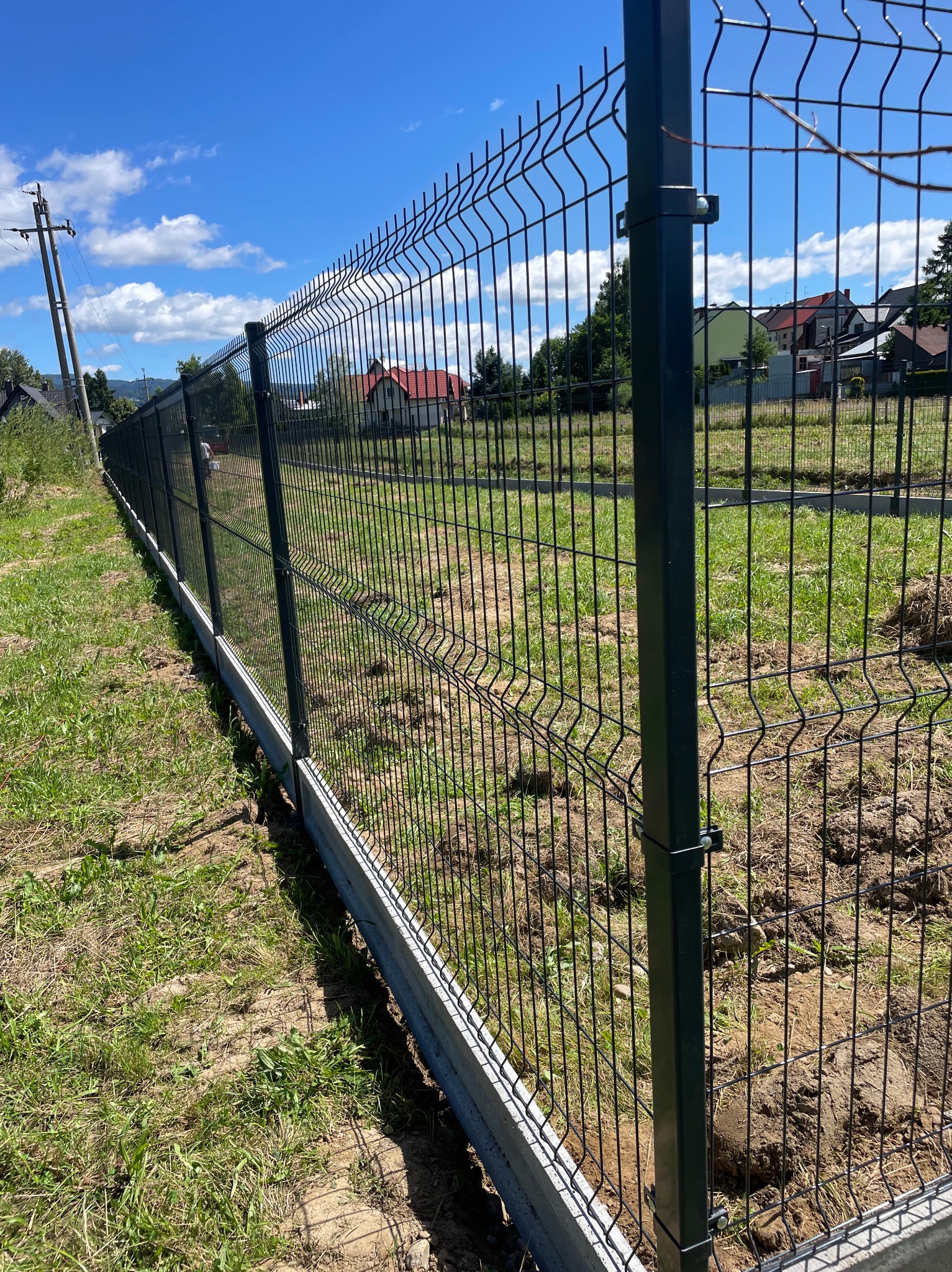 Nowoczesne, antracytowe ogrodzenie panelowe z podmurówką betonową, rozciągające się wzdłuż działki z widokiem na domy i błękitne niebo z chmurami w tle.