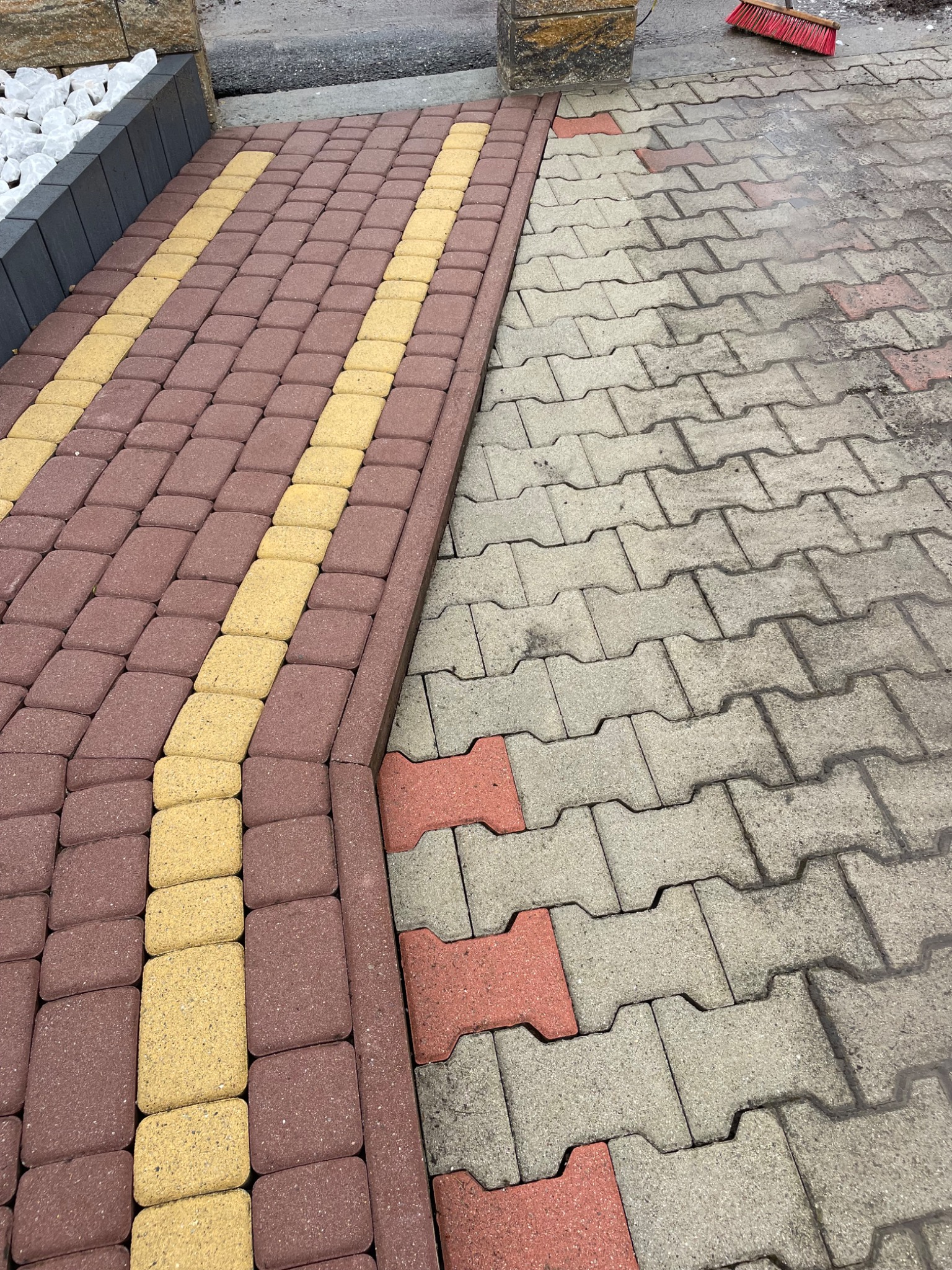 Kostka brukowa: fragment świeżo umytej powierzchni obok zabrudzonej. Widoczna czerwona szczotka. Nowa część z brązowo-żółtej kostki. Krawężnik z białymi kamieniami.