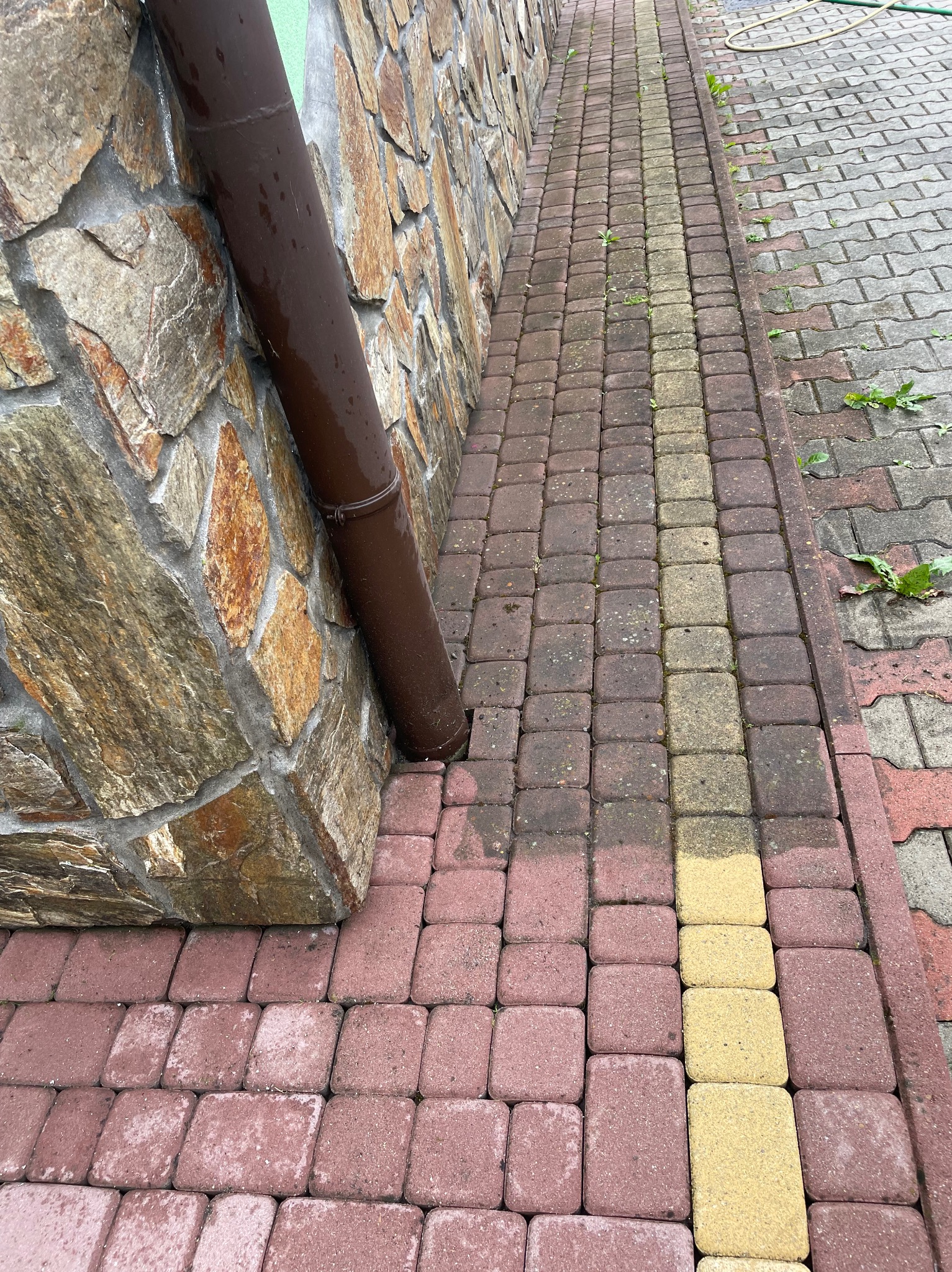 Ułożona kostka brukowa w odcieniach czerwieni i żółci, przylegająca do kamiennej ściany z brązową rynną. Widoczne zabrudzenia i chwasty na kostce.