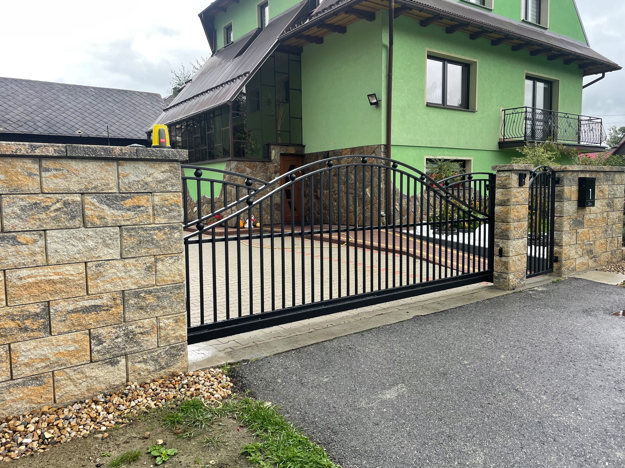 Nowoczesna, czarna brama panelowa z furtką, osadzona w murze z beżowo-brązowej kostki. W tle zielony dom z balkonem i kostka brukowa wewnątrz posesji.