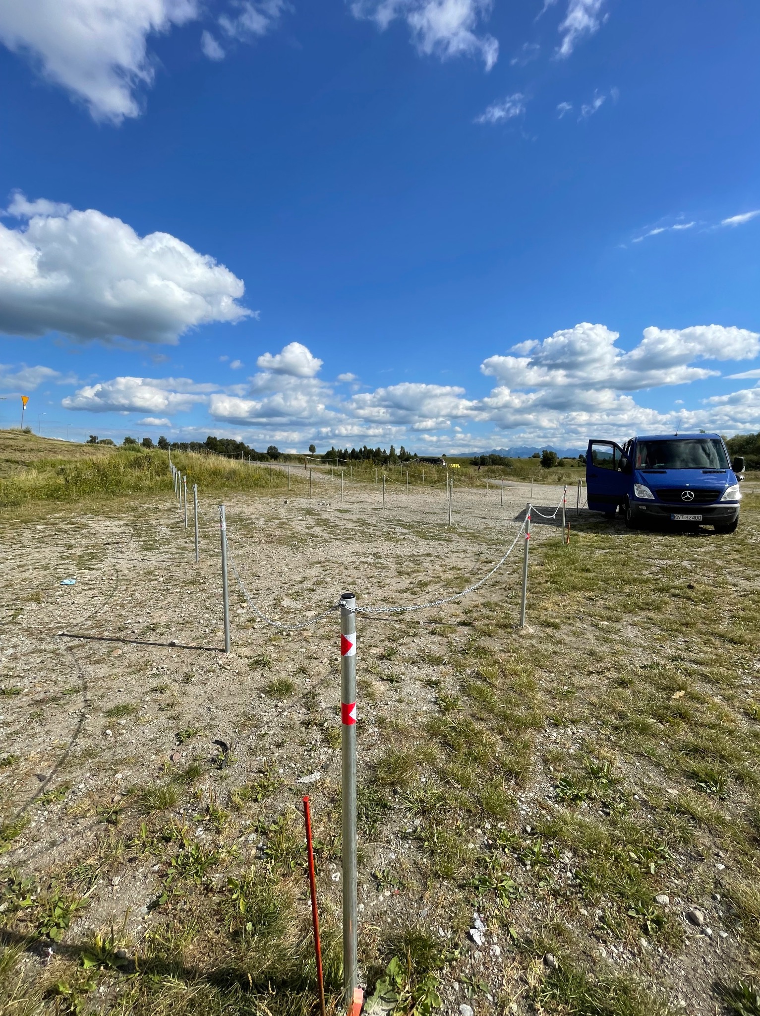 Teren wyznaczony słupkami z łańcuchem, w tle błękitny bus z otwartymi drzwiami na tle błękitnego nieba z chmurami. Etap przygotowania podłoża.