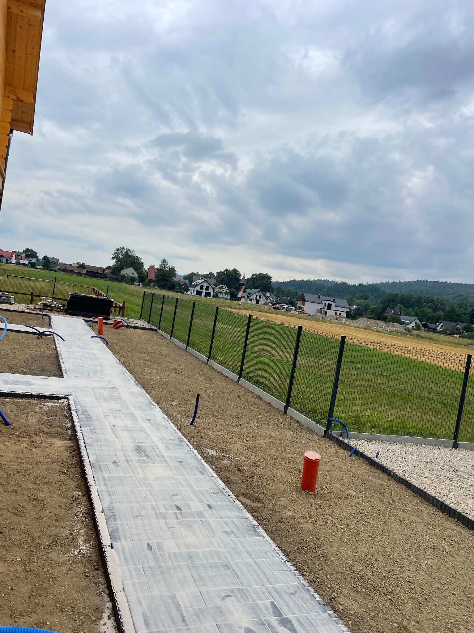 Montaż ogrodzenia panelowego i ścieżki z kostki brukowej na posesji. Widok na ułożoną ścieżkę, ogrodzenie i otaczający krajobraz. Prace w toku.