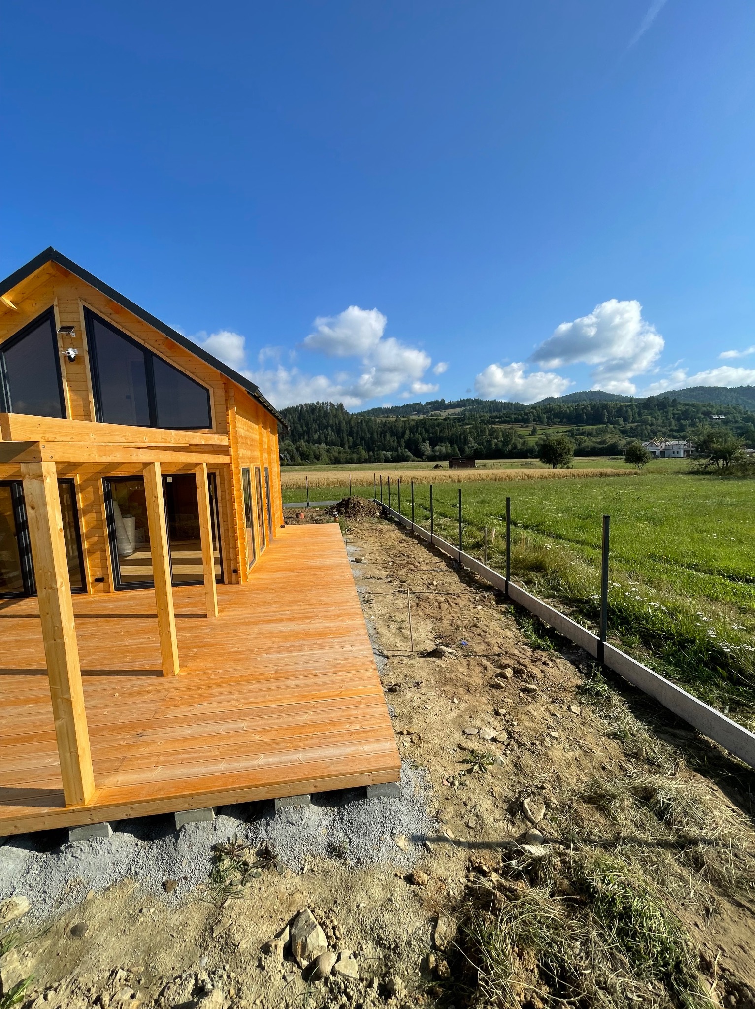 Drewniany dom z tarasem w budowie, widok na zielone wzgórza i niebo. W tle częściowo ukończone ogrodzenie panelowe. Całość w słoneczny dzień.