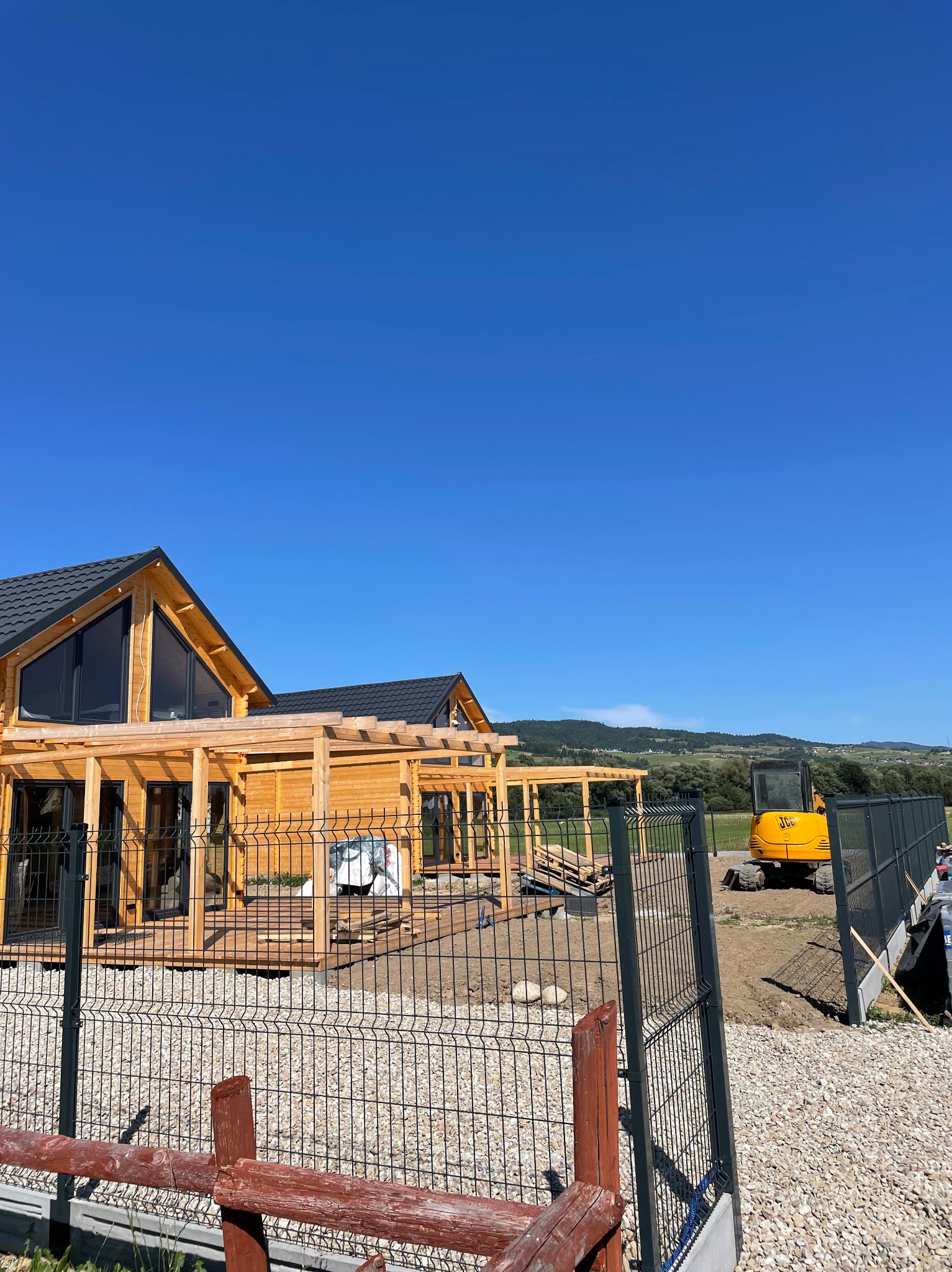 Dwa drewniane domy w trakcie budowy, z czarnymi dachami i dużymi oknami, na tle błękitnego nieba. Widoczny żółty mały traktor i fragment ogrodzenia panelowego.