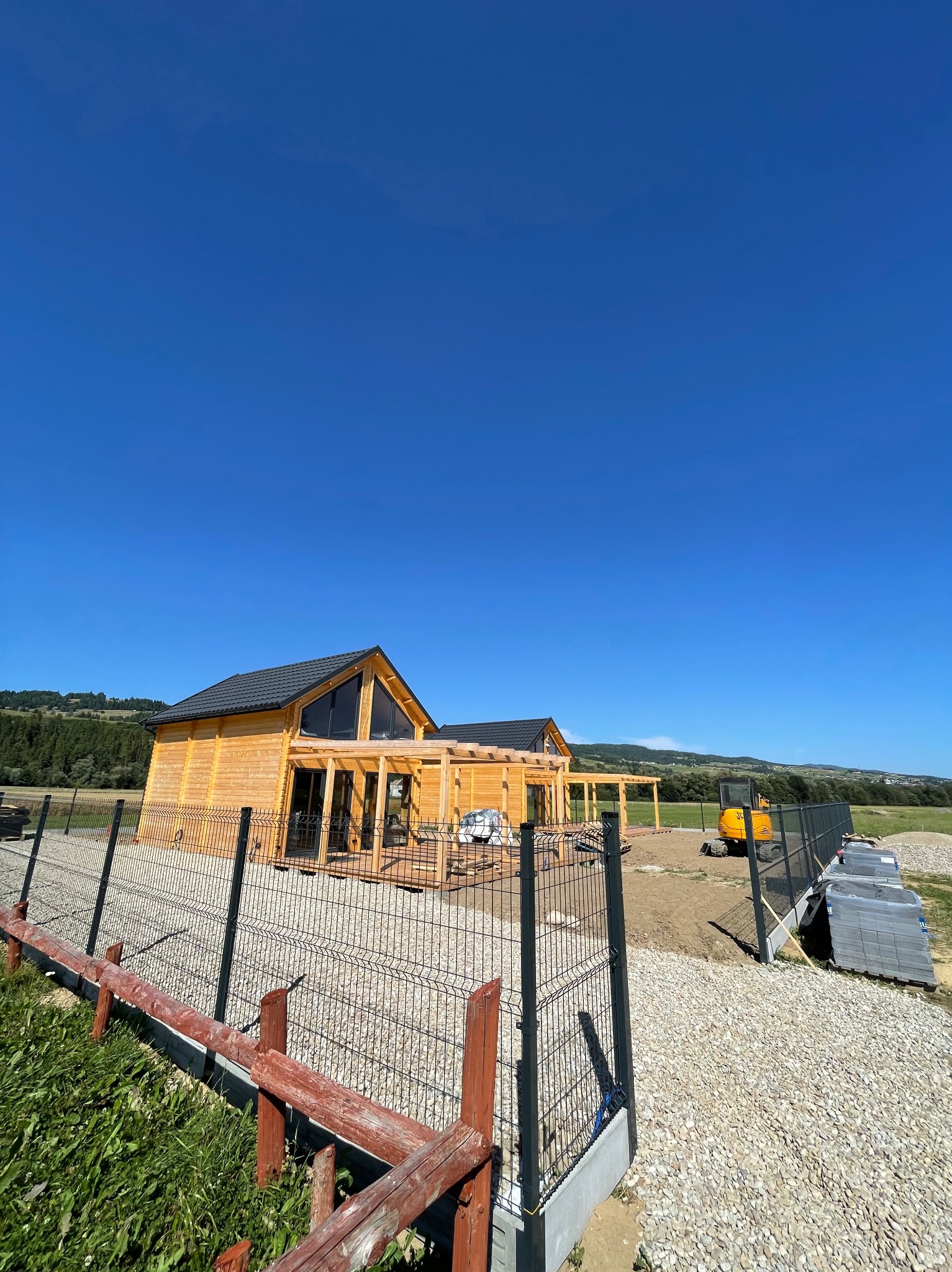 Nowoczesny, drewniany dom z czarnym dachem i dużymi oknami, otoczony ogrodzeniem panelowym i terenem w trakcie niwelacji; widoczny sprzęt budowlany.