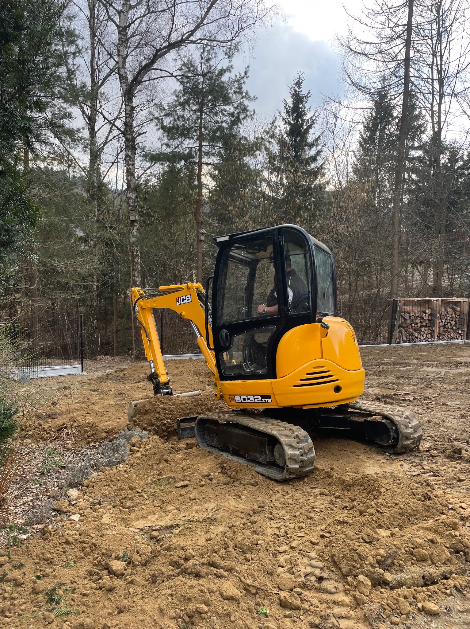 Żółta mini koparka JCB na placu budowy, w tle drzewa i częściowo widoczne ogrodzenie panelowe. Operator w kabinie. Wykopany rów w ziemi.