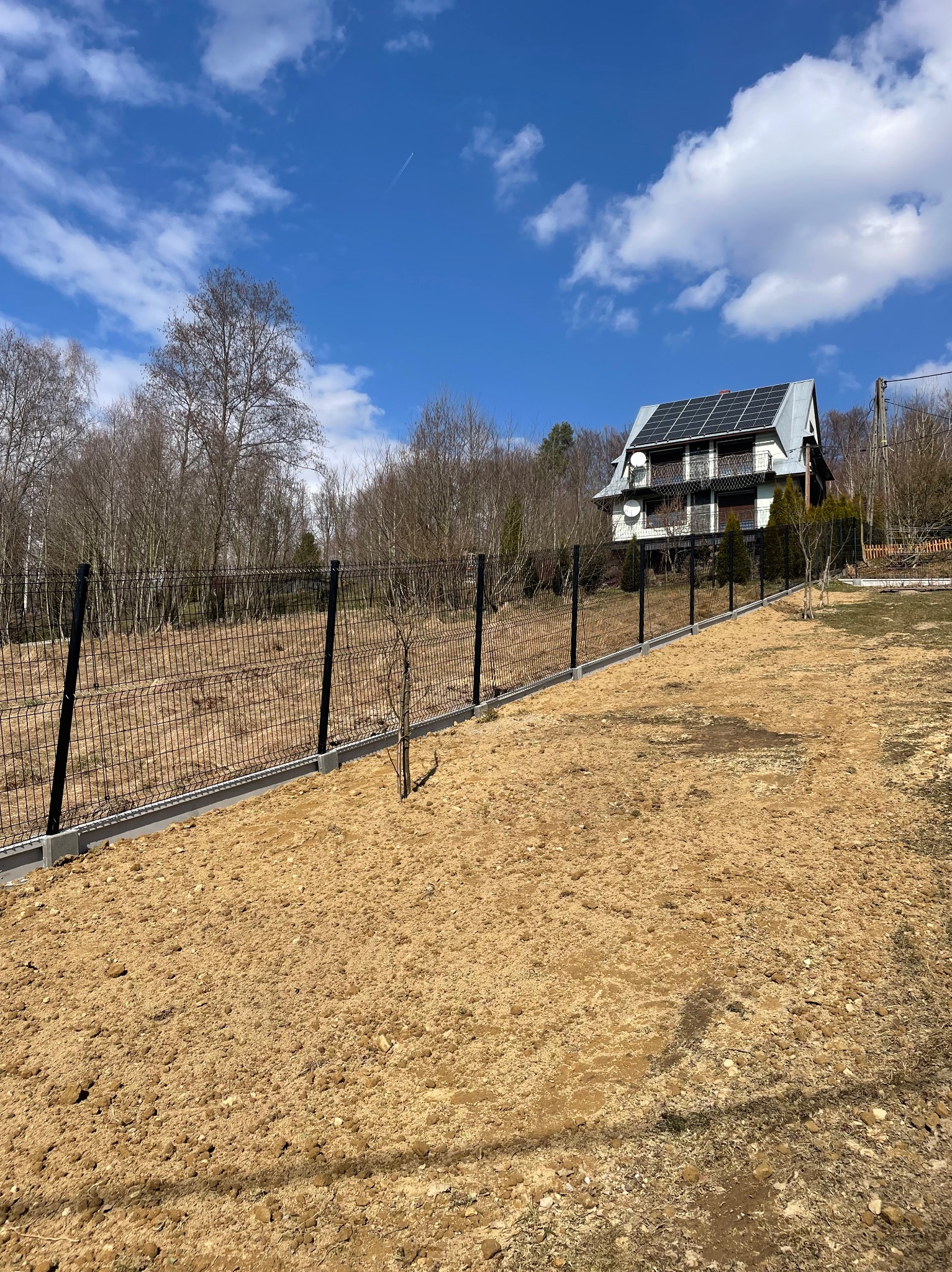 Nowoczesne ogrodzenie panelowe w kolorze czarnym, z betonową podmurówką, otaczające posesję z domem z panelami słonecznymi w tle, na przygotowanym terenie.
