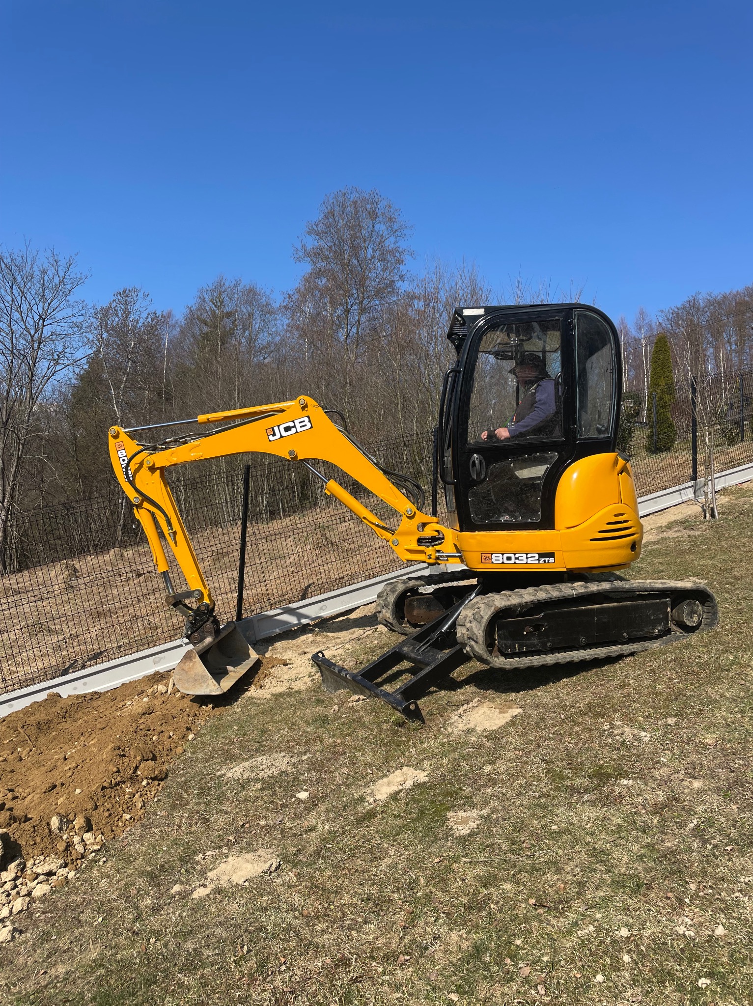 Żółta mini koparka JCB 8032 ZTS w trakcie prac ziemnych przy nowym ogrodzeniu panelowym na trawiastym terenie w słoneczny dzień. Operator w kabinie.