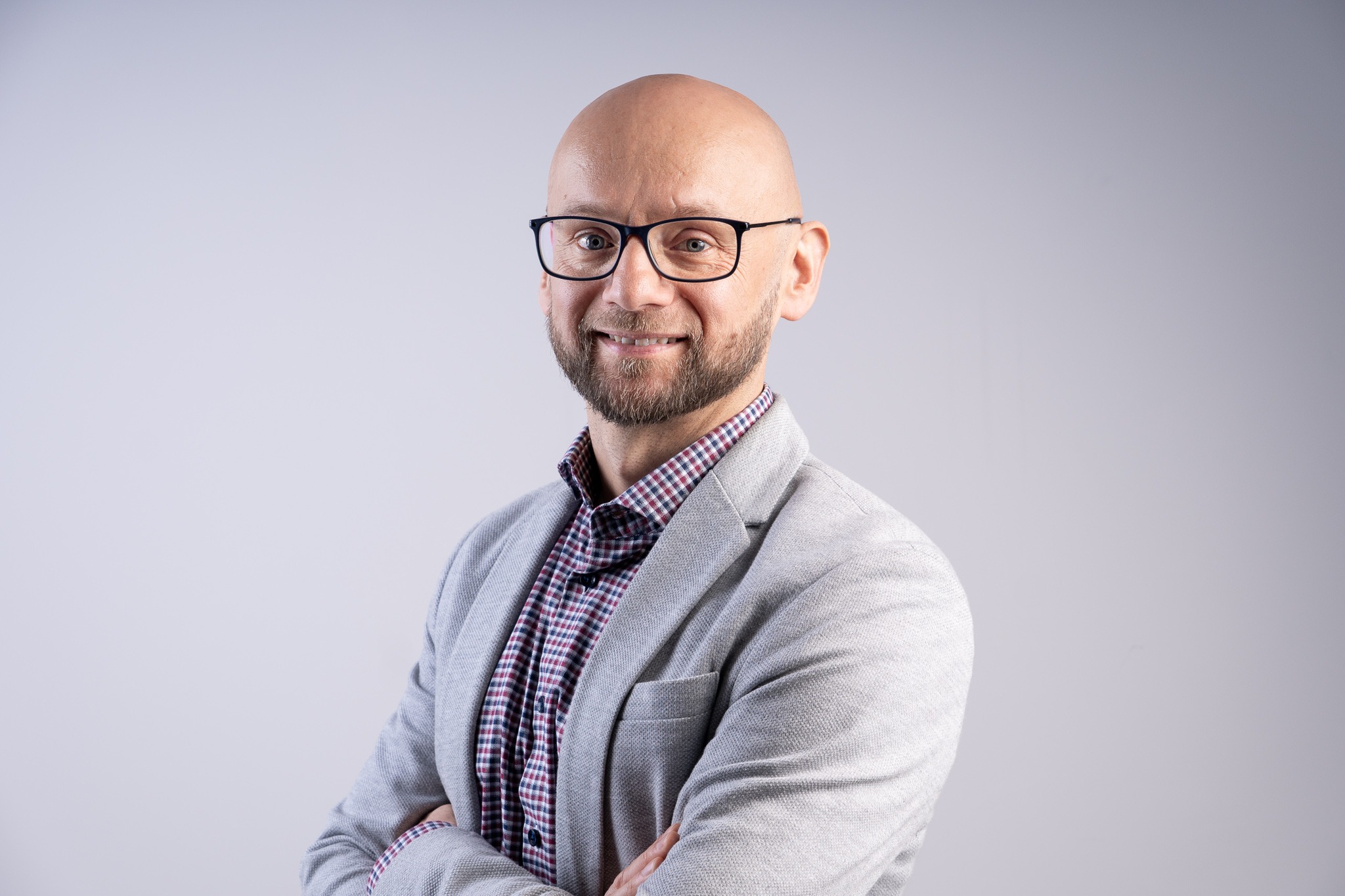 Portret uśmiechniętego mężczyzny w okularach, z brodą, ubranego w szarą marynarkę i koszulę w kratę, na jednolitym jasnym tle.