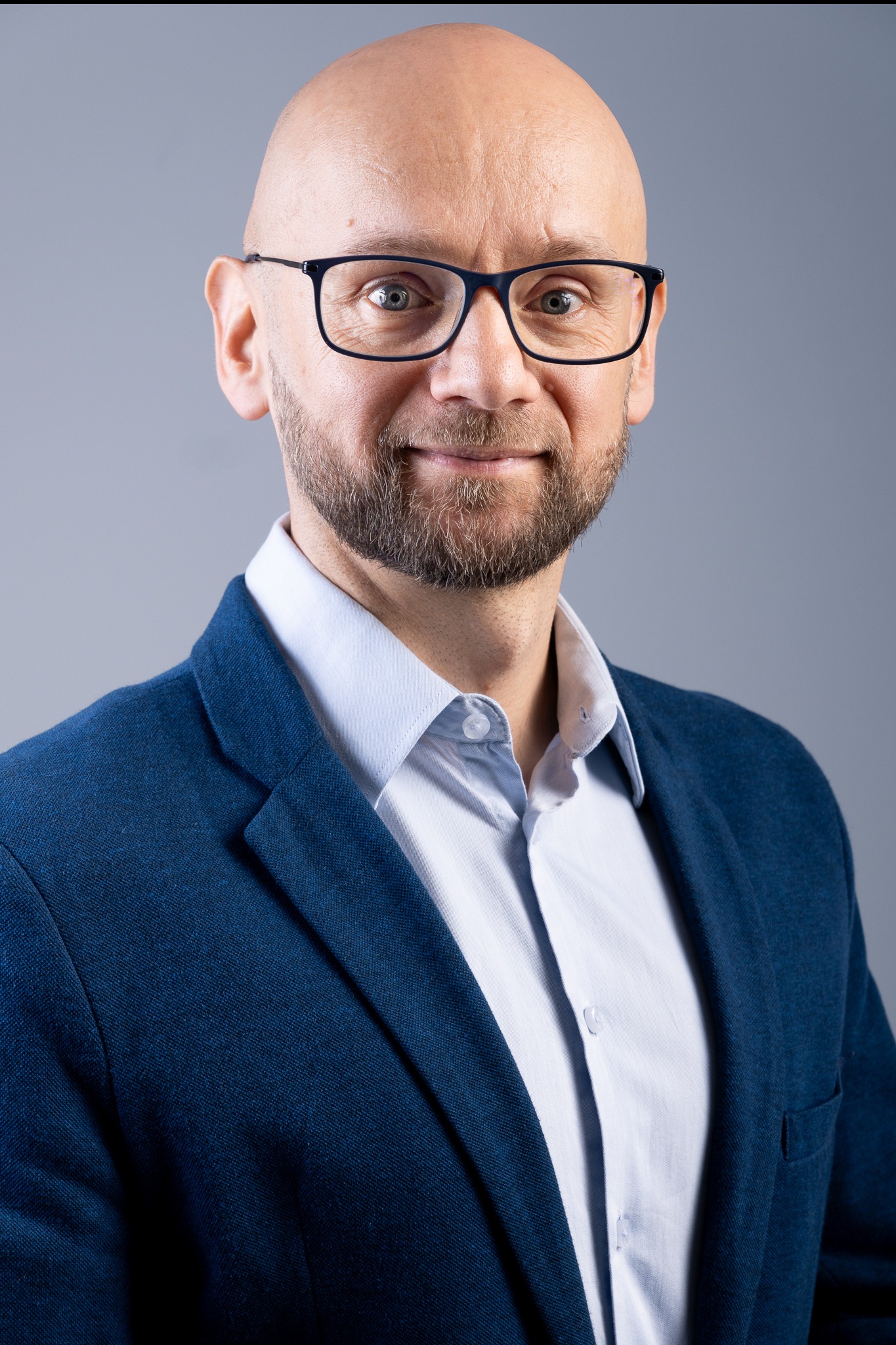Portret uśmiechniętego, łysiejącego mężczyzny w okularach, ubrany w niebieską marynarkę i jasnoniebieską koszulę, na jednolitym szarym tle.