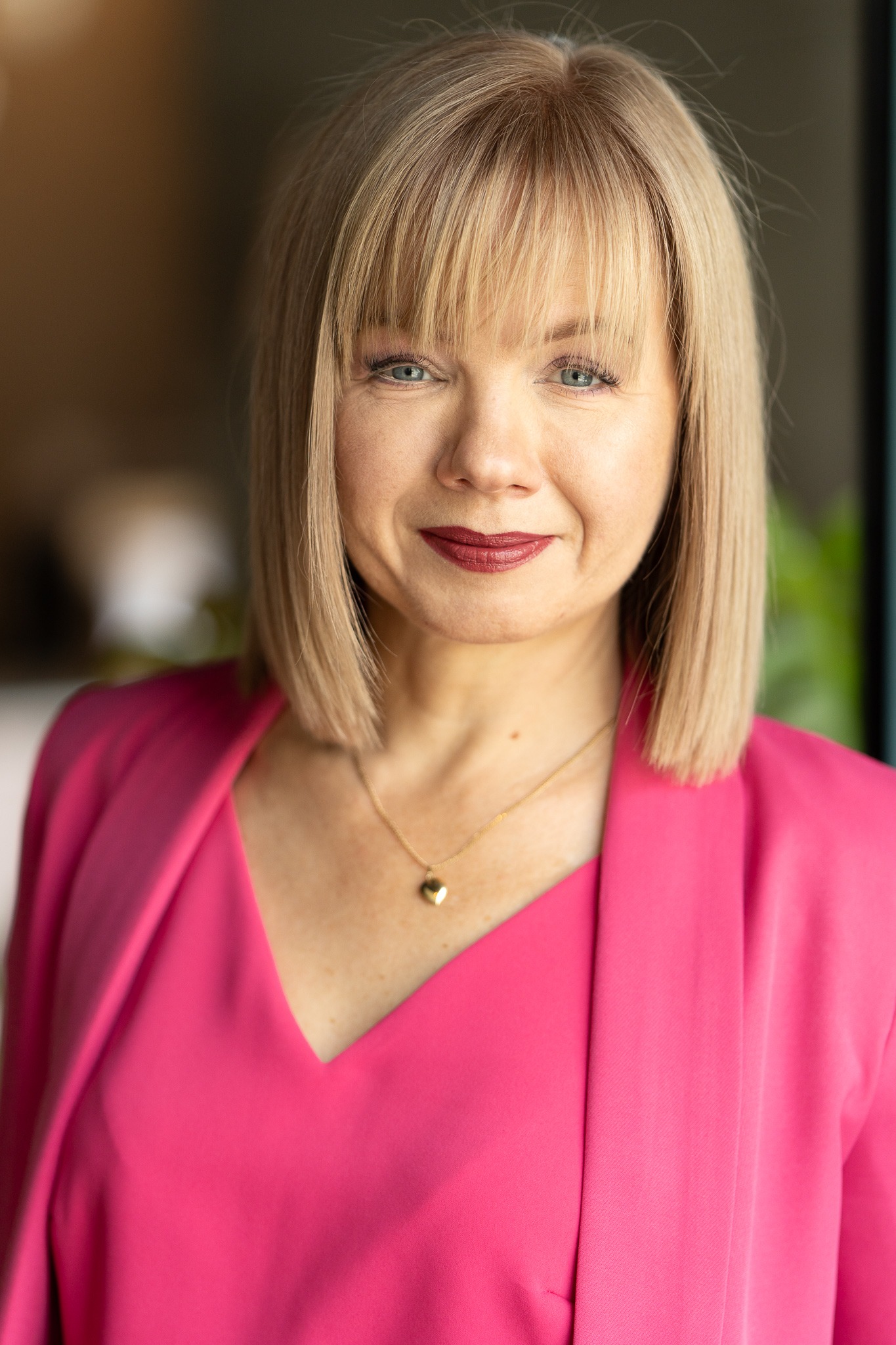 Portret kobiety z blond bobem i grzywką, ubranej w różowy żakiet, z delikatnym złotym naszyjnikiem w kształcie serca, patrzącej w obiektyw z lekkim uśmiechem.