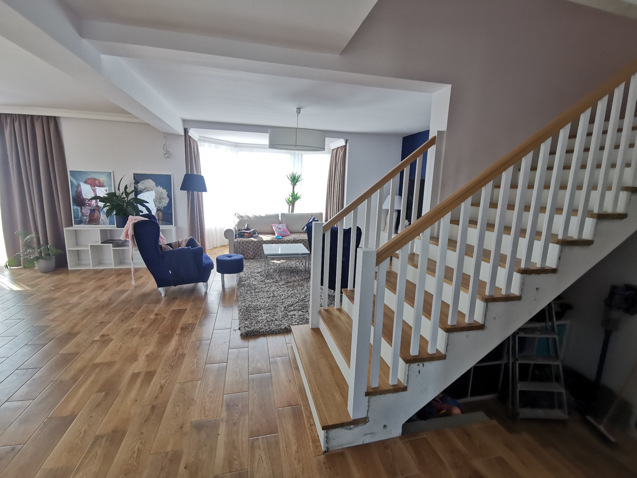 Widok na wnętrze domu z drewnianą podłogą, schodami z białymi balustradami i drewnianymi stopniami, salon z niebieskim fotelem i dywanem w tle.