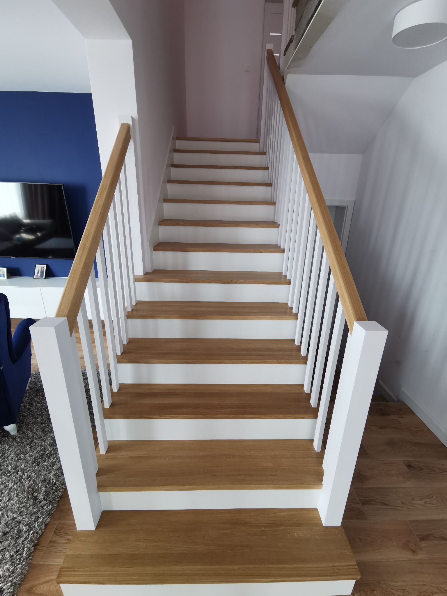 Proste schody z drewnianymi stopniami i białymi podstopnicami oraz balustradą z drewnianą poręczą, widok z dołu w górę.