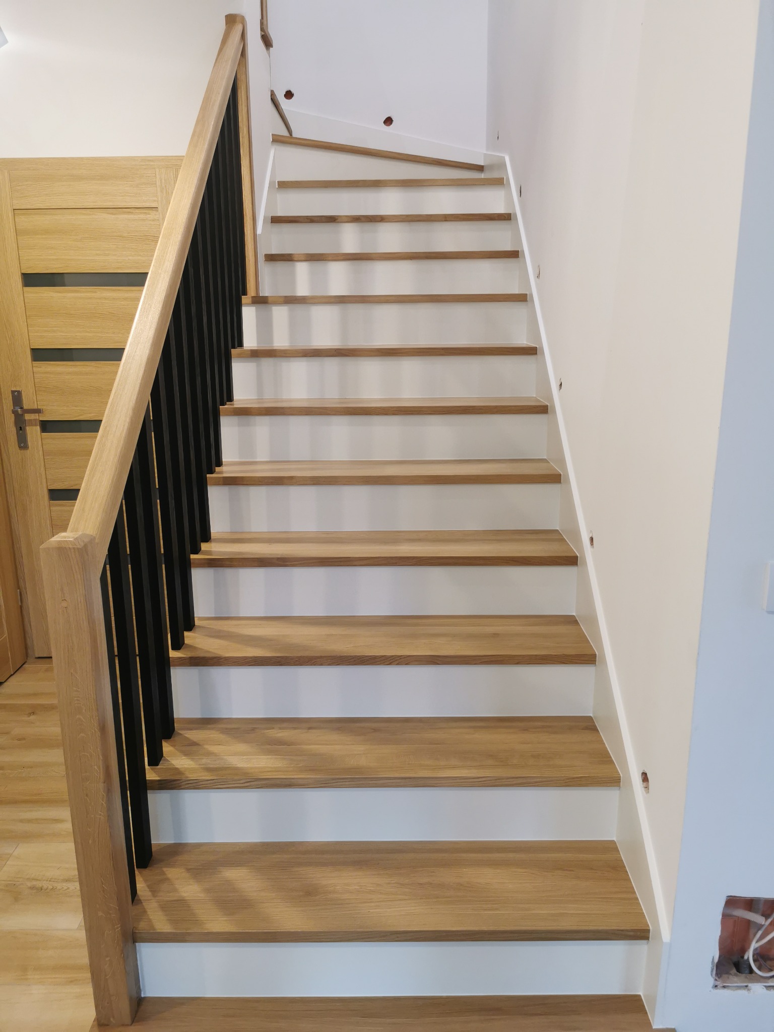 Proste schody z drewnianymi stopniami i białą podstopnicą, z czarną balustradą i drewnianą poręczą, widok z dołu.