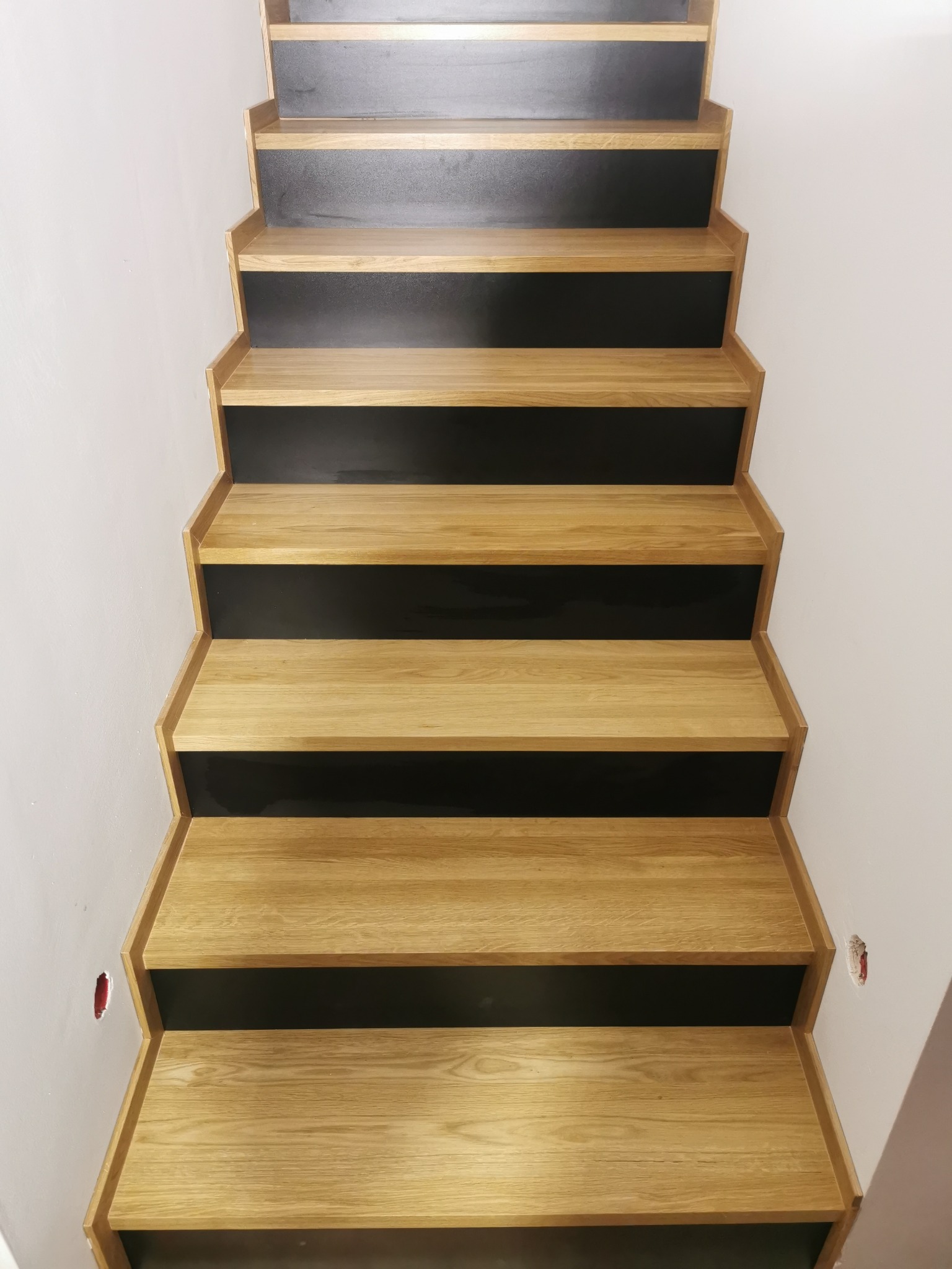 Widok z dołu na schody drewniane z czarnymi podstopnicami, widoczne niedokończone wykończenie ściany obok.