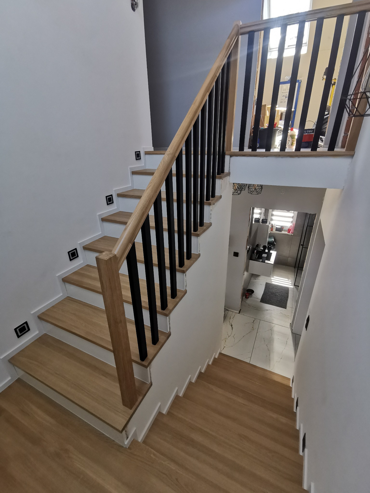 Nowoczesne schody z drewnianymi stopniami i czarną balustradą, oświetlenie punktowe na ścianie, widok na marmurową podłogę w holu.